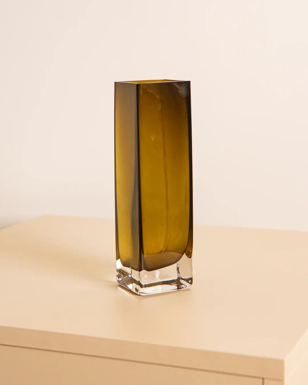A tall, rectangular amber Murano glass vase from the 80s collection by Treaptyque sits on a beige surface against a light-colored wall, exuding an air of refined elegance.