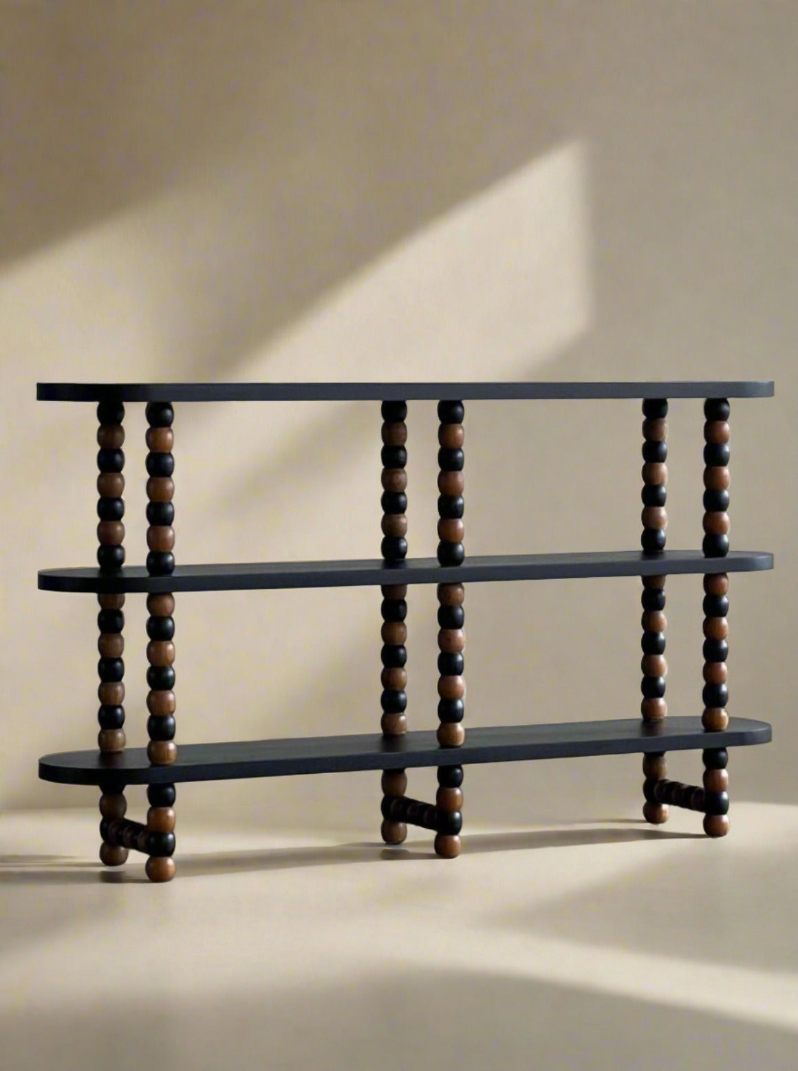 A modern three-tiered wood shelf stands against a plain wall. The shelves are supported by vertical columns made of alternating black and natural wood spheres, creating beaded supports. The sleek, minimalist design of the Studio Kallang Onde Shelf contrasts with the concrete floor below.