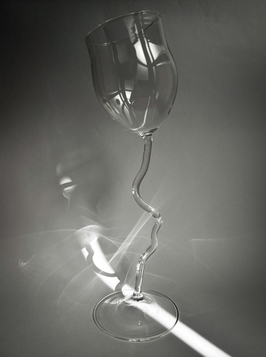 The Agustina Ros Long Wine Glass features a wavy, twisted stem and stands on a flat surface, casting abstract reflections and shadows in the light against a gray background.