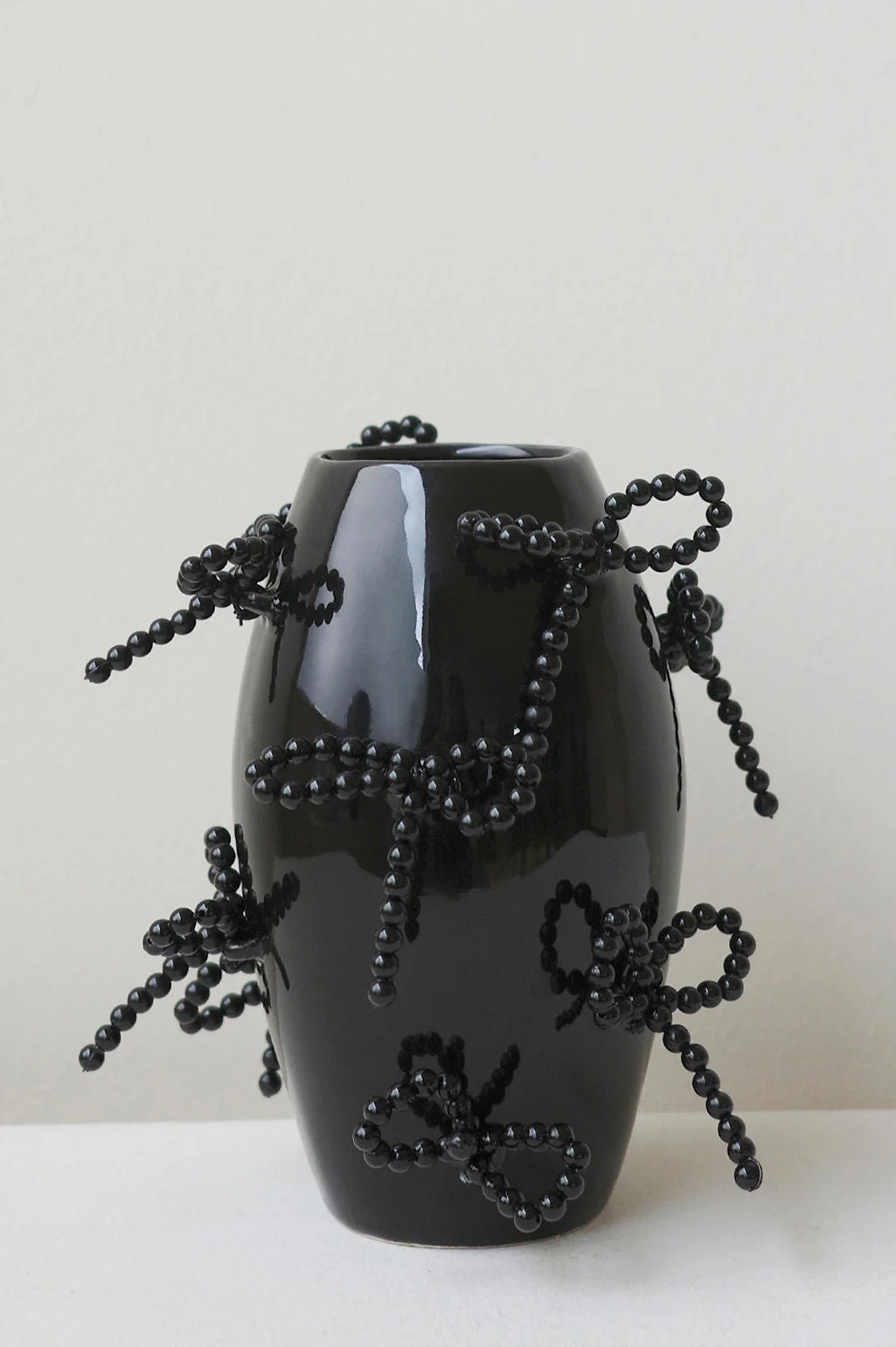 The Pearly Pearl small vase in gloss black by Completedworks boasts a sculptural form with abstract beaded bows. Its smooth, reflective surface elegantly fuses modern design with traditional ceramics against a light gray backdrop.