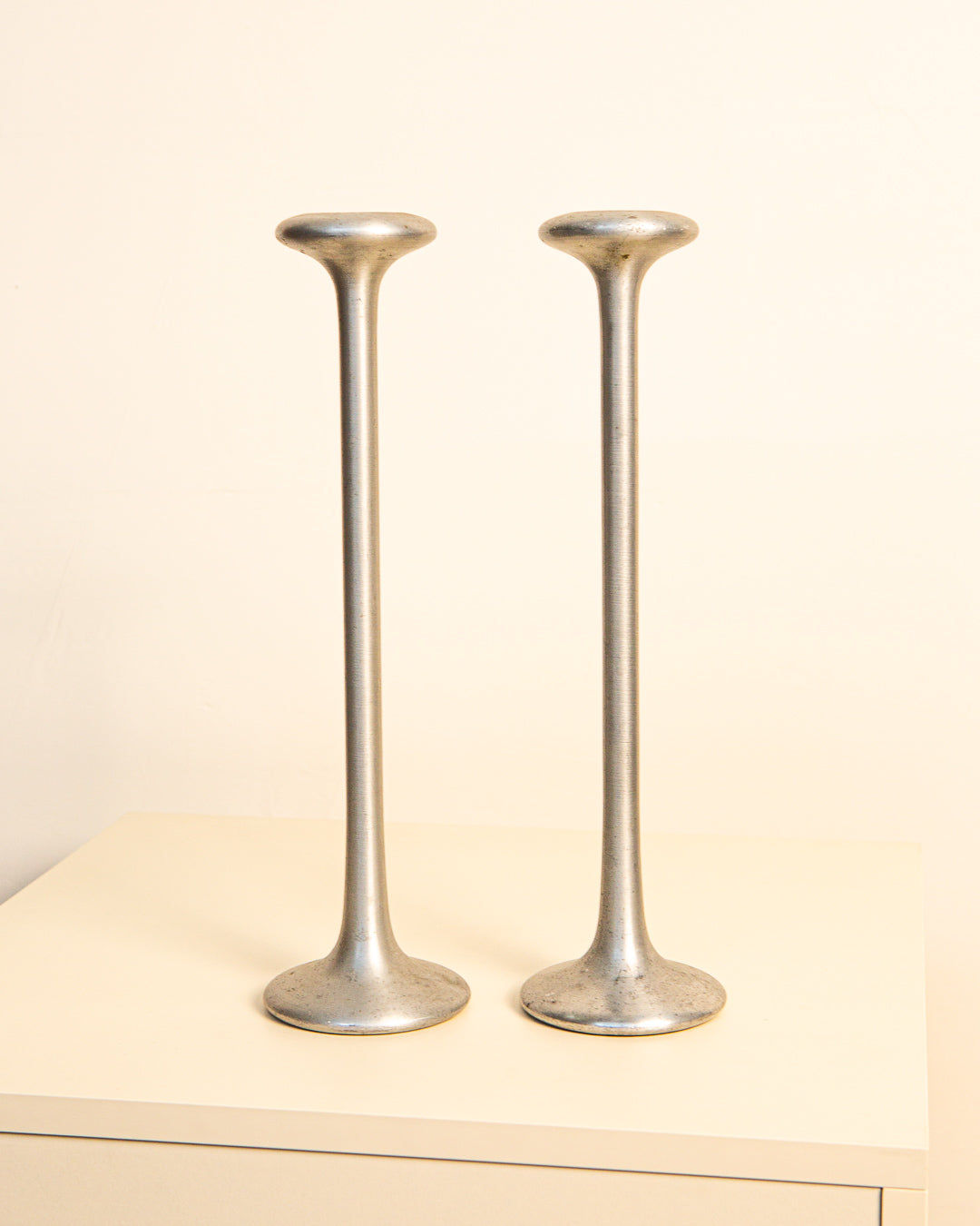 A Pair of Cast Aluminum Candleholders by Treaptyque, featuring tall, slender forms with wide bases and rounded tops, are displayed side by side on a light surface, highlighting their minimalist 1980s industrial design.