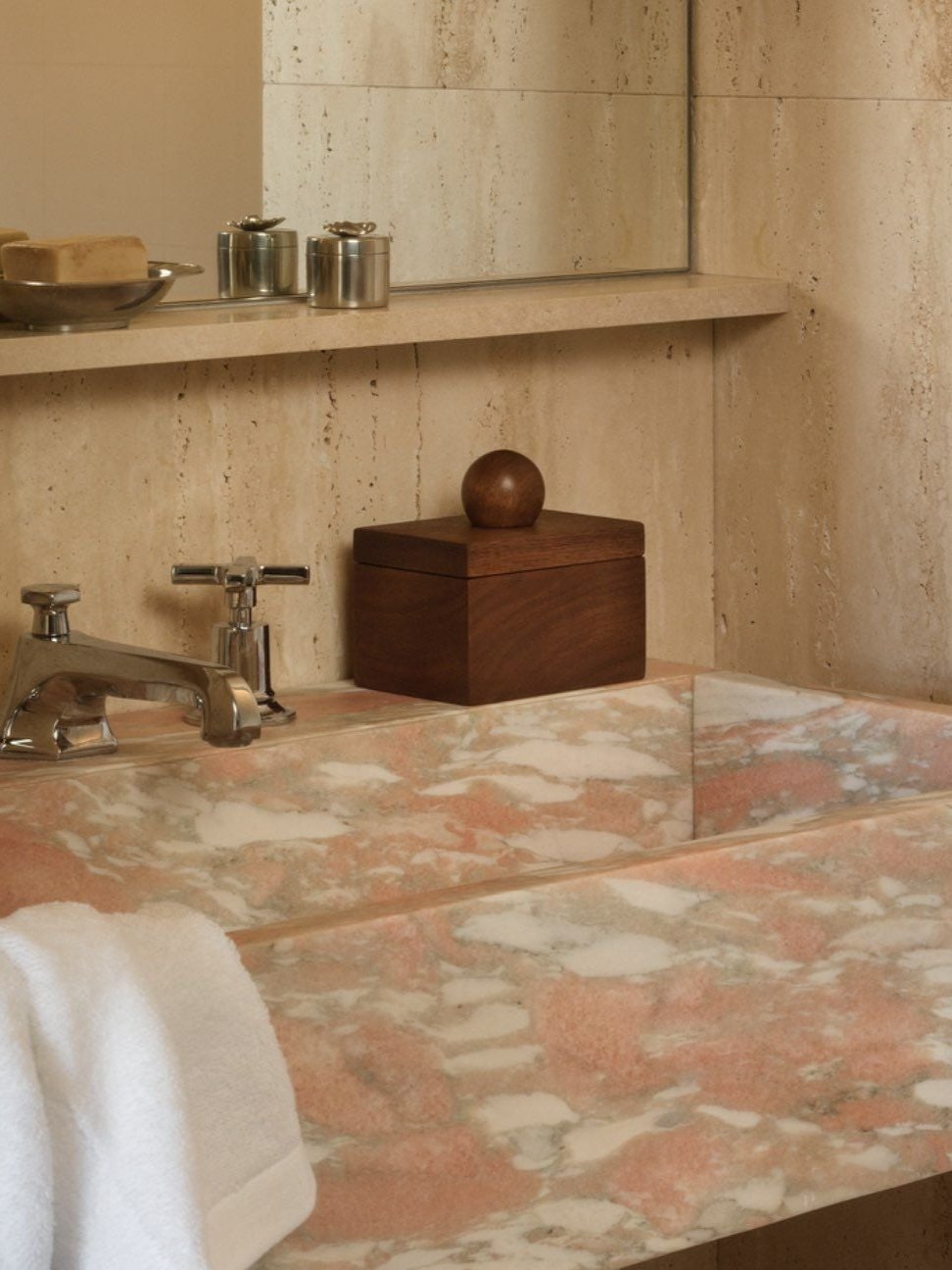 A stylish bathroom showcases a rectangular marble sink with shades of pink and white, accentuating its geometric lines. A white towel is draped over the edge, while above, a shelf holds soap alongside Studio Vraco's Box 01 Walnut. A large mirror reflects the elegant stone walls.