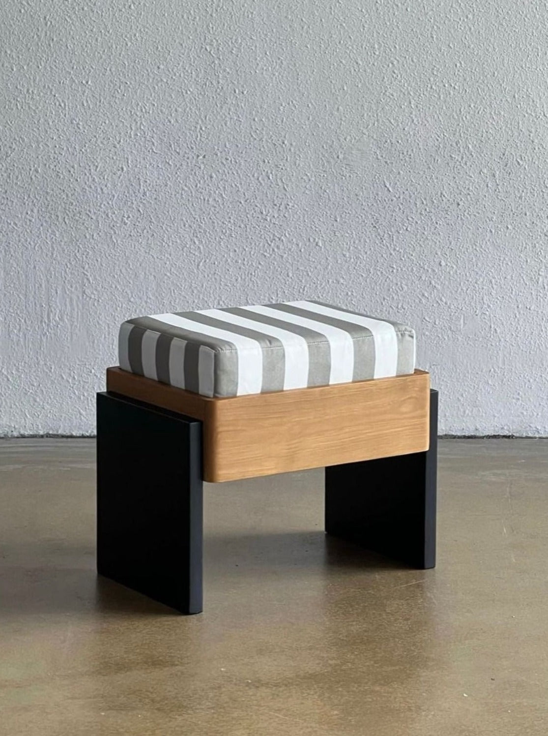 A modern Striped Ottoman by Studio Kallang featuring a rectangular wooden base with black legs and a striped cushion on top. The cushion has a white and gray pattern. The ottoman is placed on a polished concrete floor against a plain light gray wall.