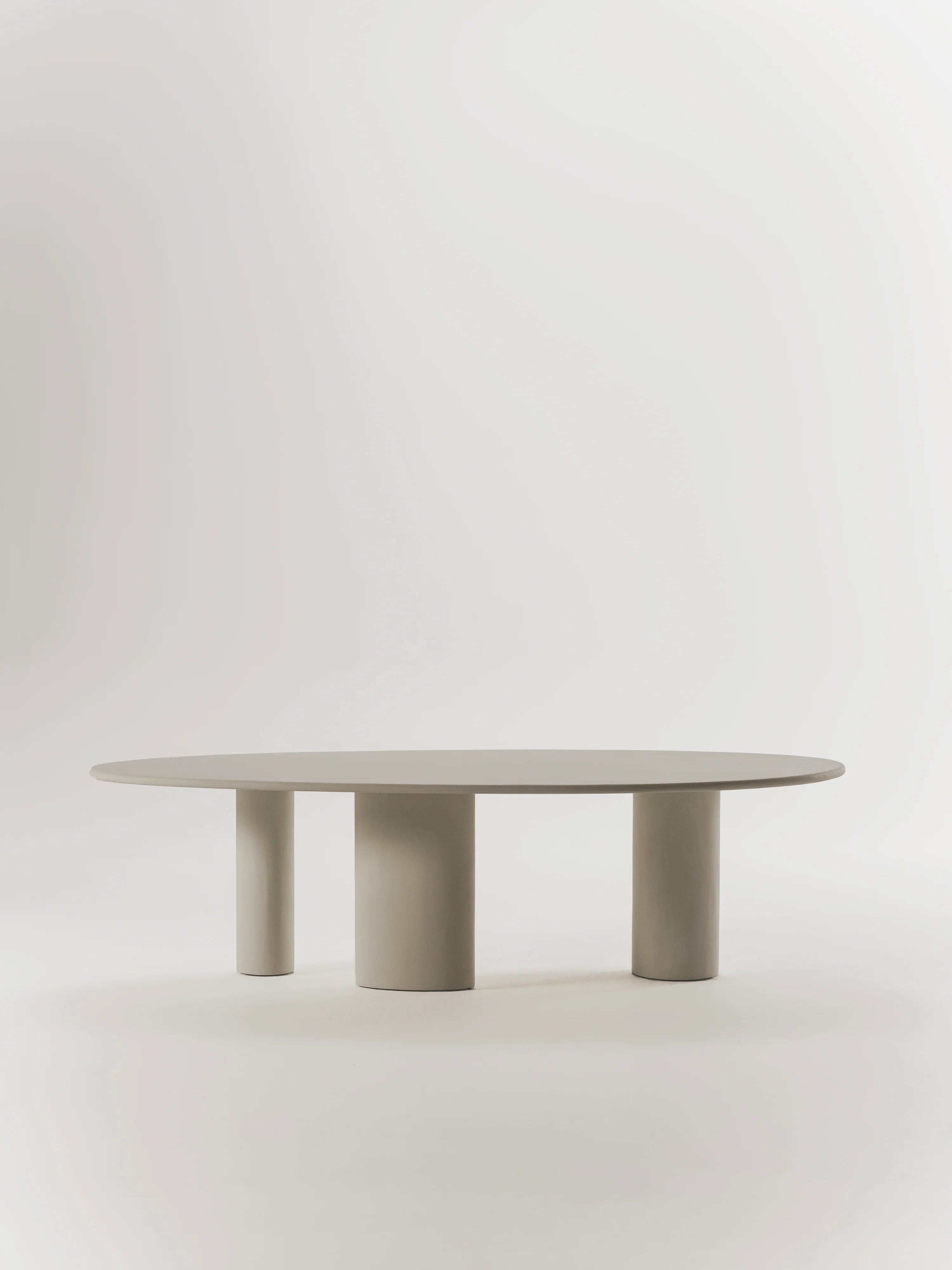 The "Graça" Dining Table by MarlotBaus is a minimalist design featuring a round top and three cylindrical legs, handcrafted in neutral beige microcement, set against a plain light background.