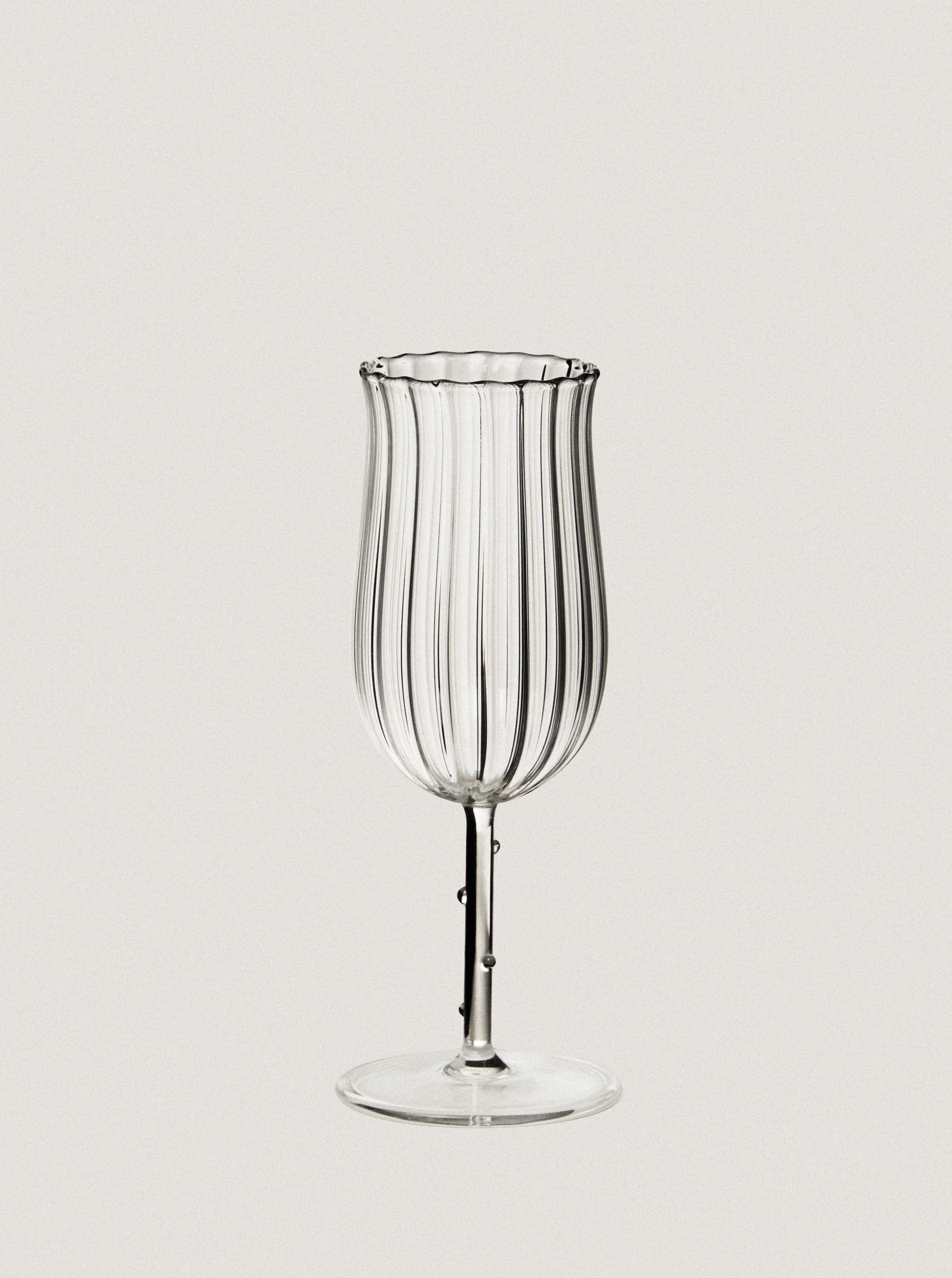 A clear, vertically striped Tulip Wine Glass - Set of Four by Sophie Lou Jacobsen with a slightly fluted rim stands against a plain off-white background. The glass features a slender stem and a circular base, designed to enhance the release of aromas.