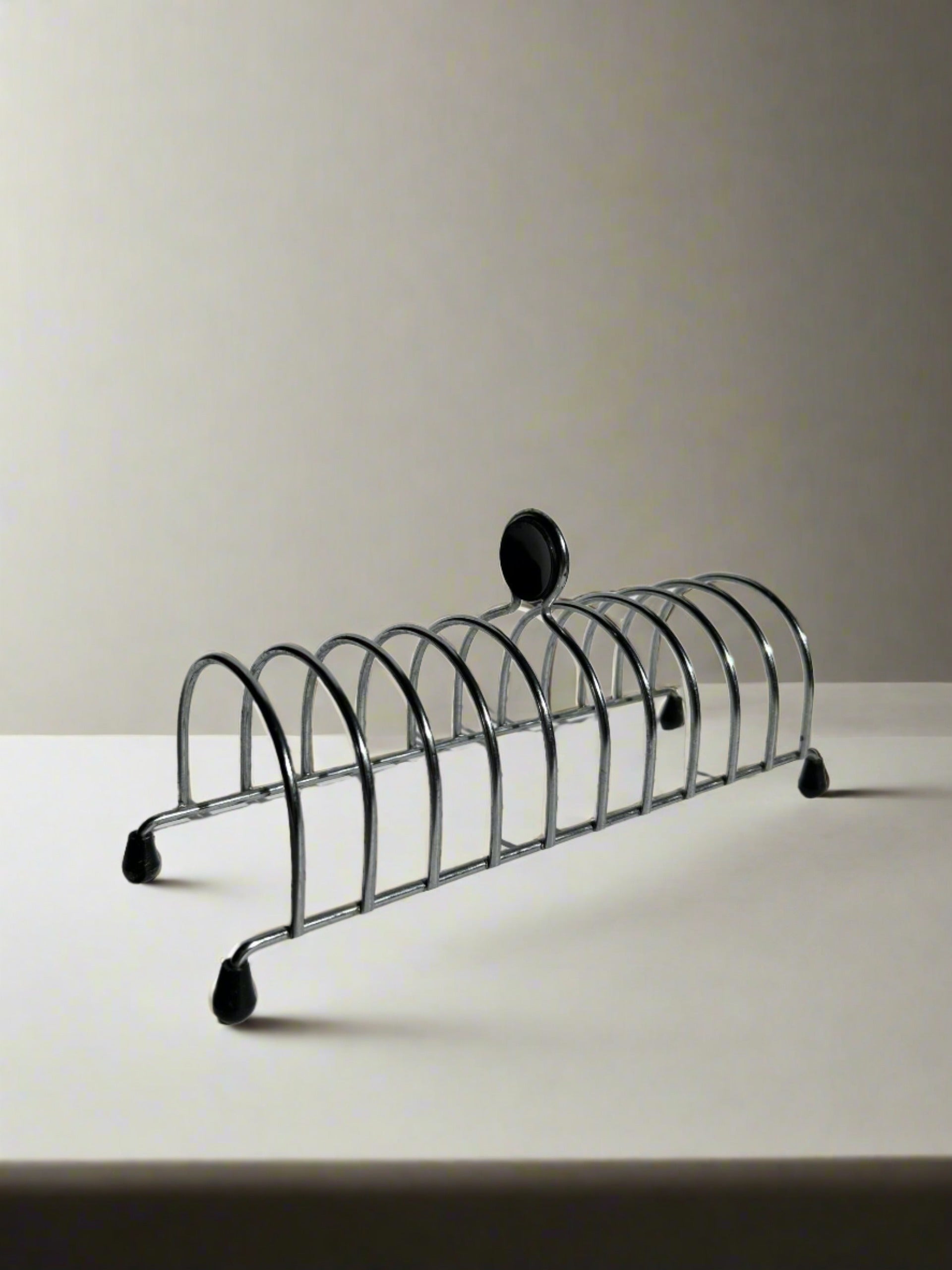 The Postmodern Toast Rack from Flock Studio, designed by Tomado Holland, evokes a 1950s style with multiple slots to hold toast upright on a white tabletop. It showcases a neutral background and is finished with a small black knob.