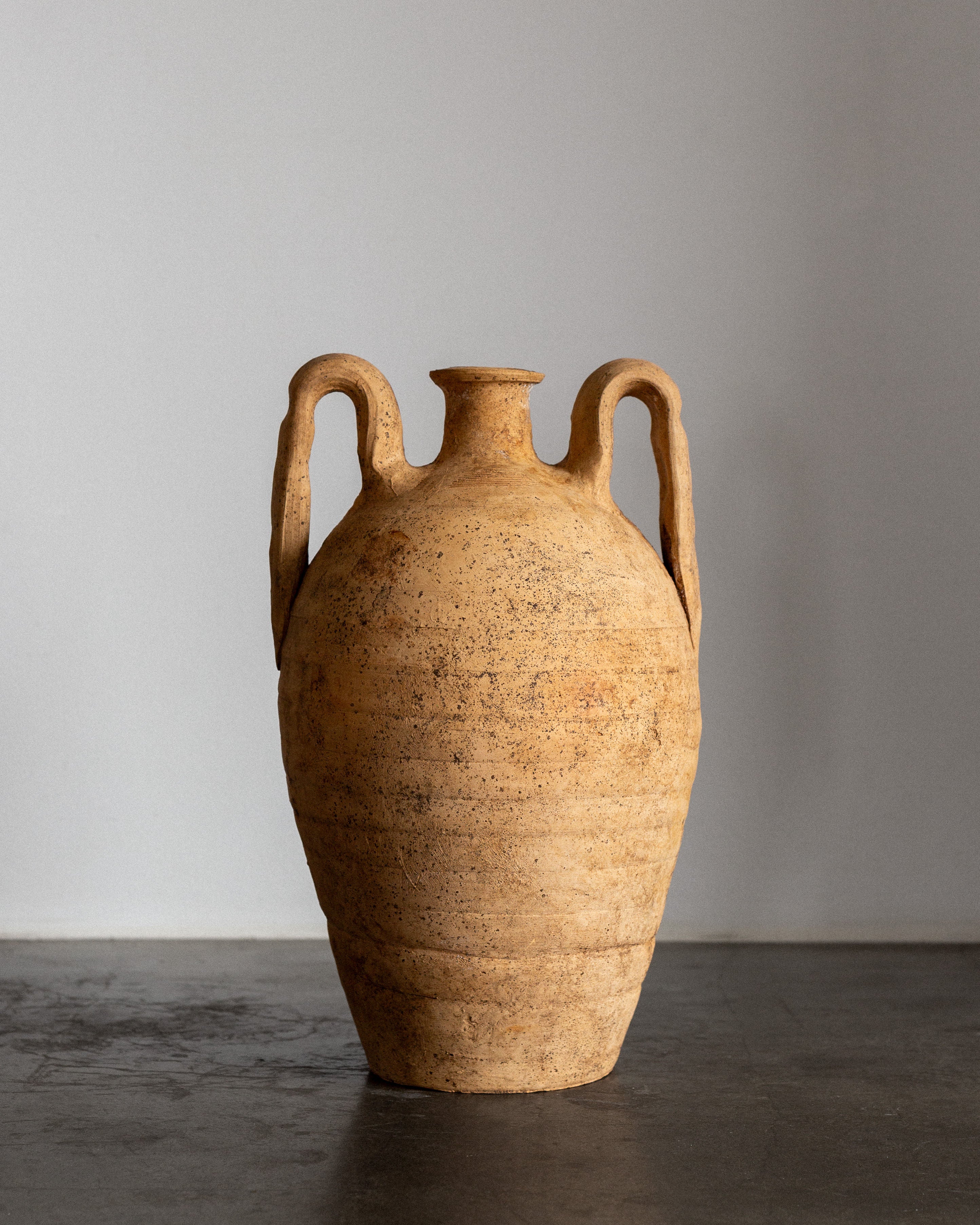 The Early 20th Century European Ceramic Amphora by Spigel, a large beige earthenware piece with textured, speckled finish and wide side handles, rests on a smooth dark surface against a light gray background, making it an exquisite decorative item.
