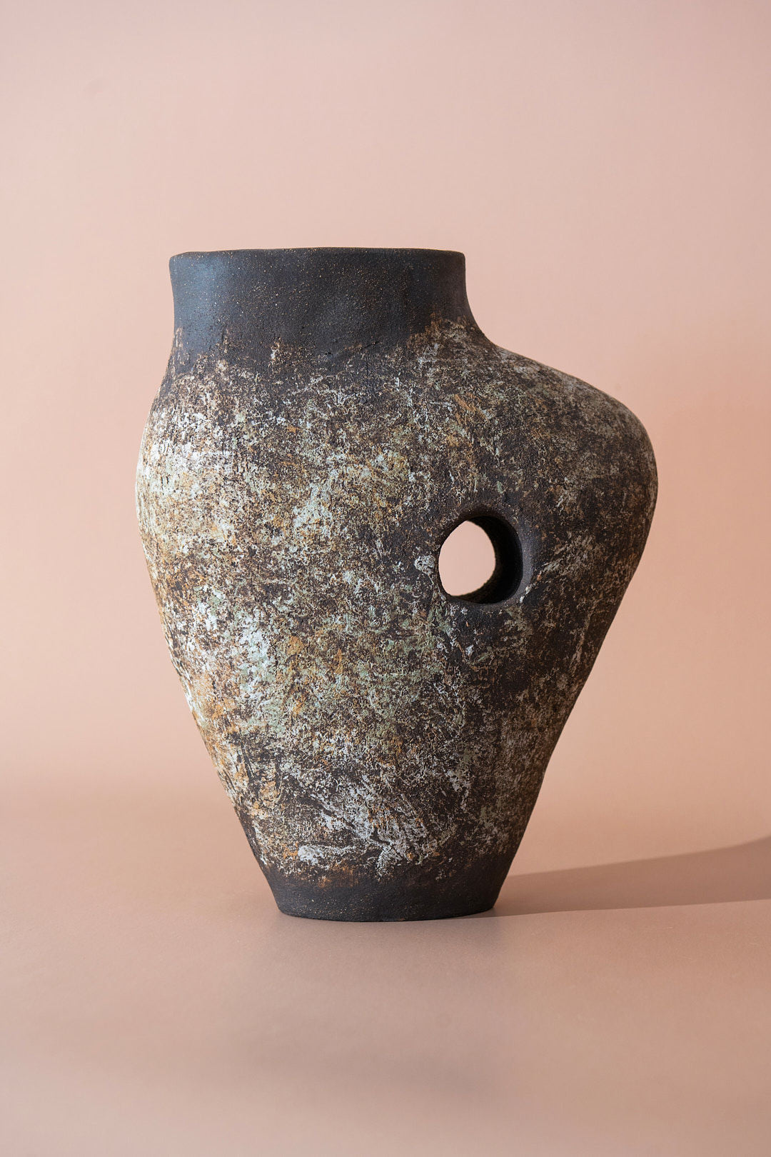 The Silhouette Vase 06 - The Tunnel Series by Siham Djebbar Ceramics is a rustic, asymmetrical stoneware vase with an earthy texture and a small circular hole, set against a neutral beige background.