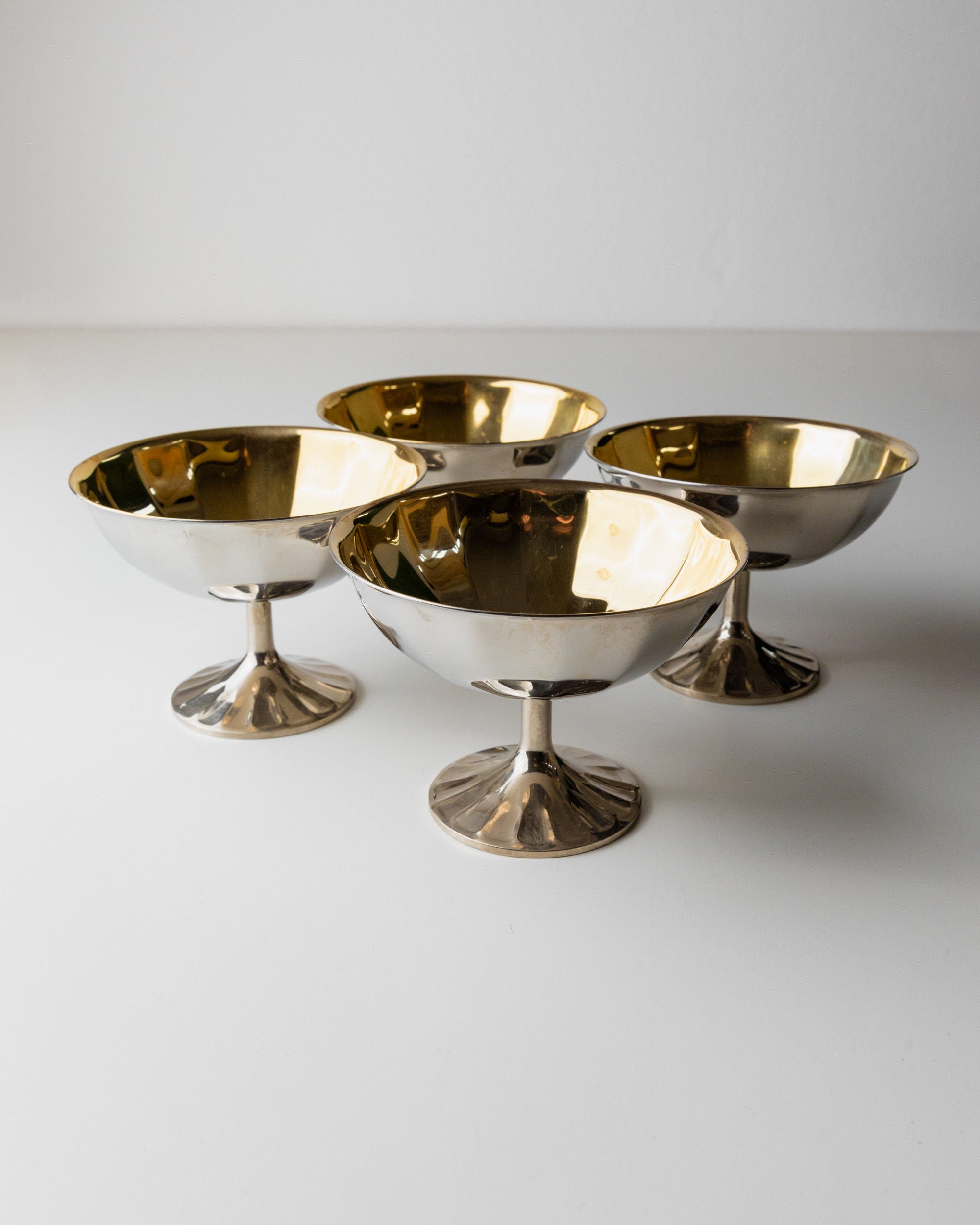 Four Champagne Coupes by Bottega Jacobs, crafted in Italy in the 1970s with a shimmering blend of silver-plated and gold-toned metal, stand elegantly on a white surface. Their reflective finish and graceful stems catch the light, showcasing their ornate metallic texture.