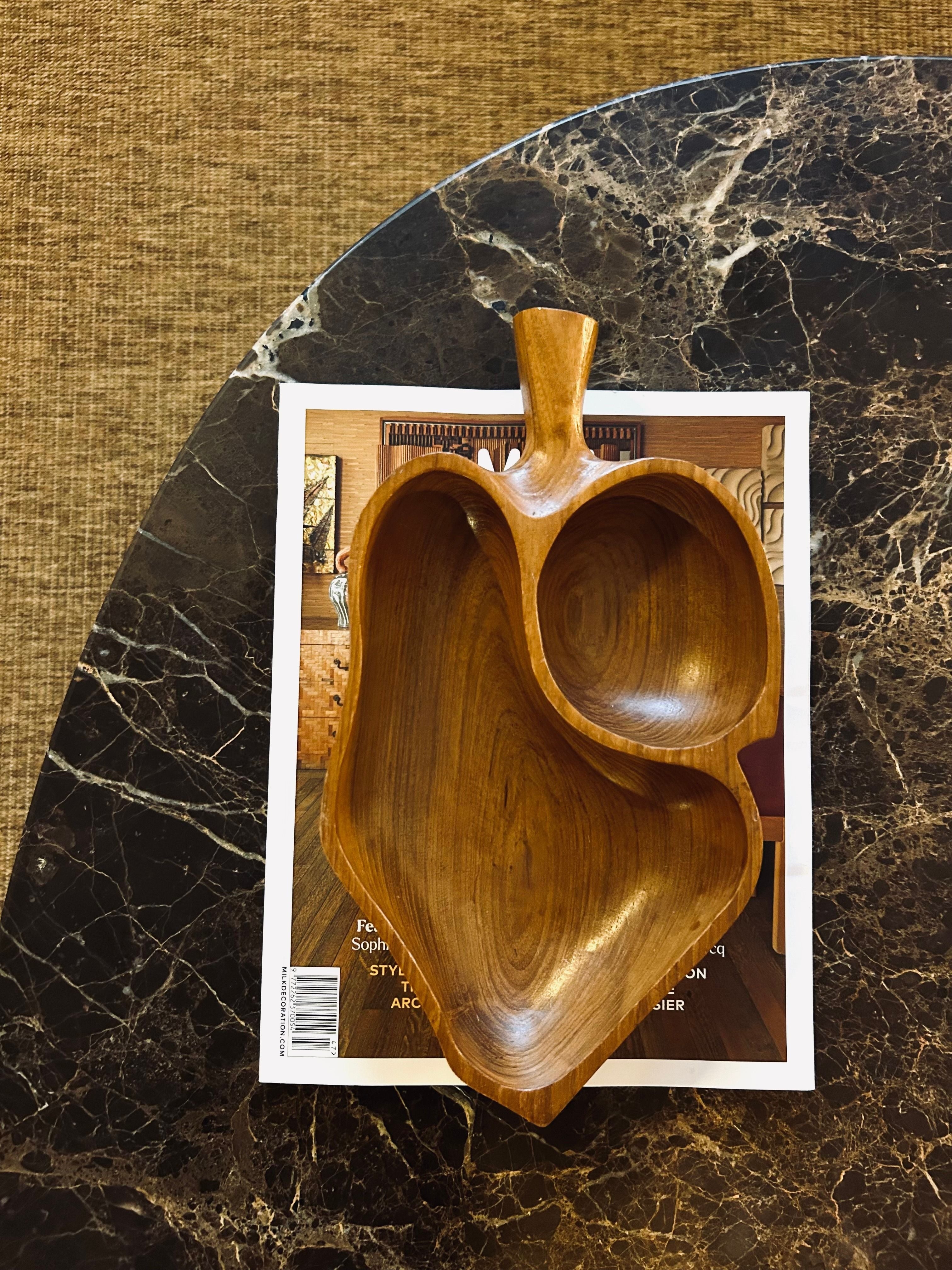 A Danish Teak Leaf Bowl by Flock Studio, featuring two compartments and embodying Scandinavian design, is elegantly displayed on an open magazine. The magazine lies atop a dark, textured marble surface, accentuating the bowl's inherent durability and sophistication.