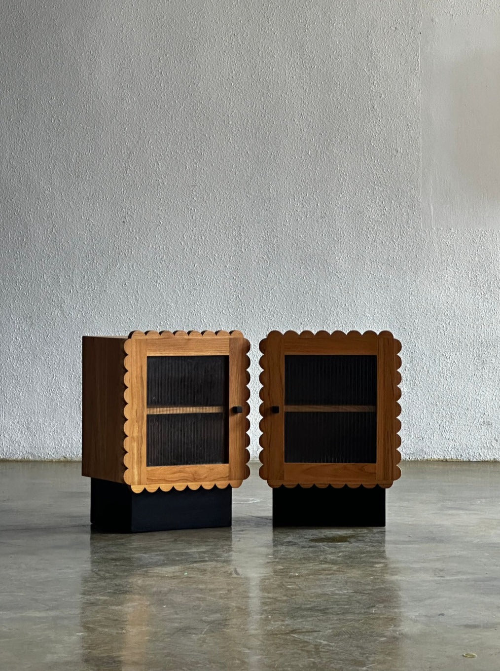 A pair of Mamun No. 2 Night Stands by Studio Kallang with scalloped edges and black base, featuring fluted glass-front doors, are positioned on a polished concrete floor against a plain wall. The modern design blends rustic elements with contemporary style.
