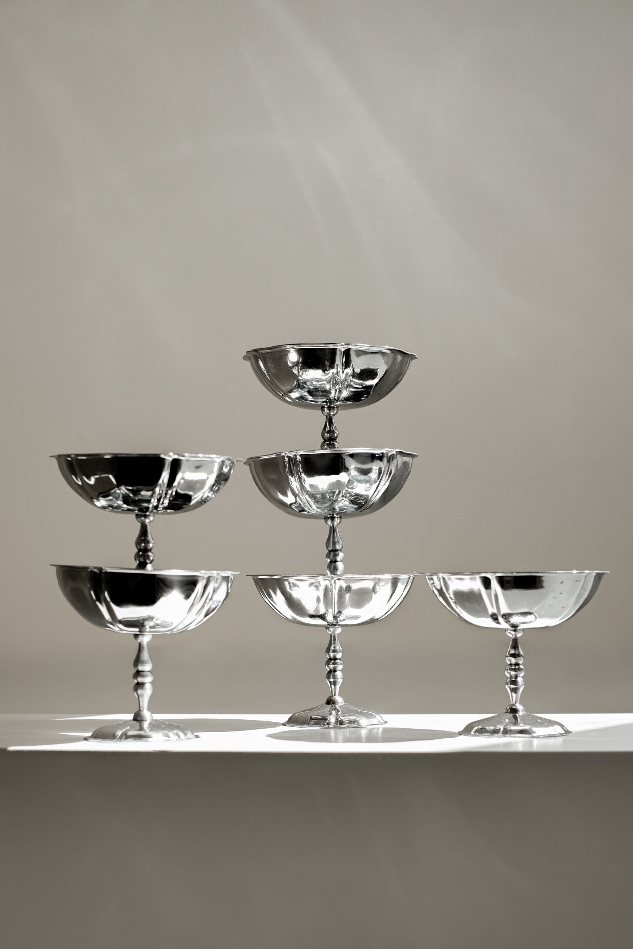 The Set of Six Ice Coupes by septembre studios, stacked in a pyramid on a white surface, showcases stainless steel shine and vintage appeal with subtle reflections and shadows against a plain background.