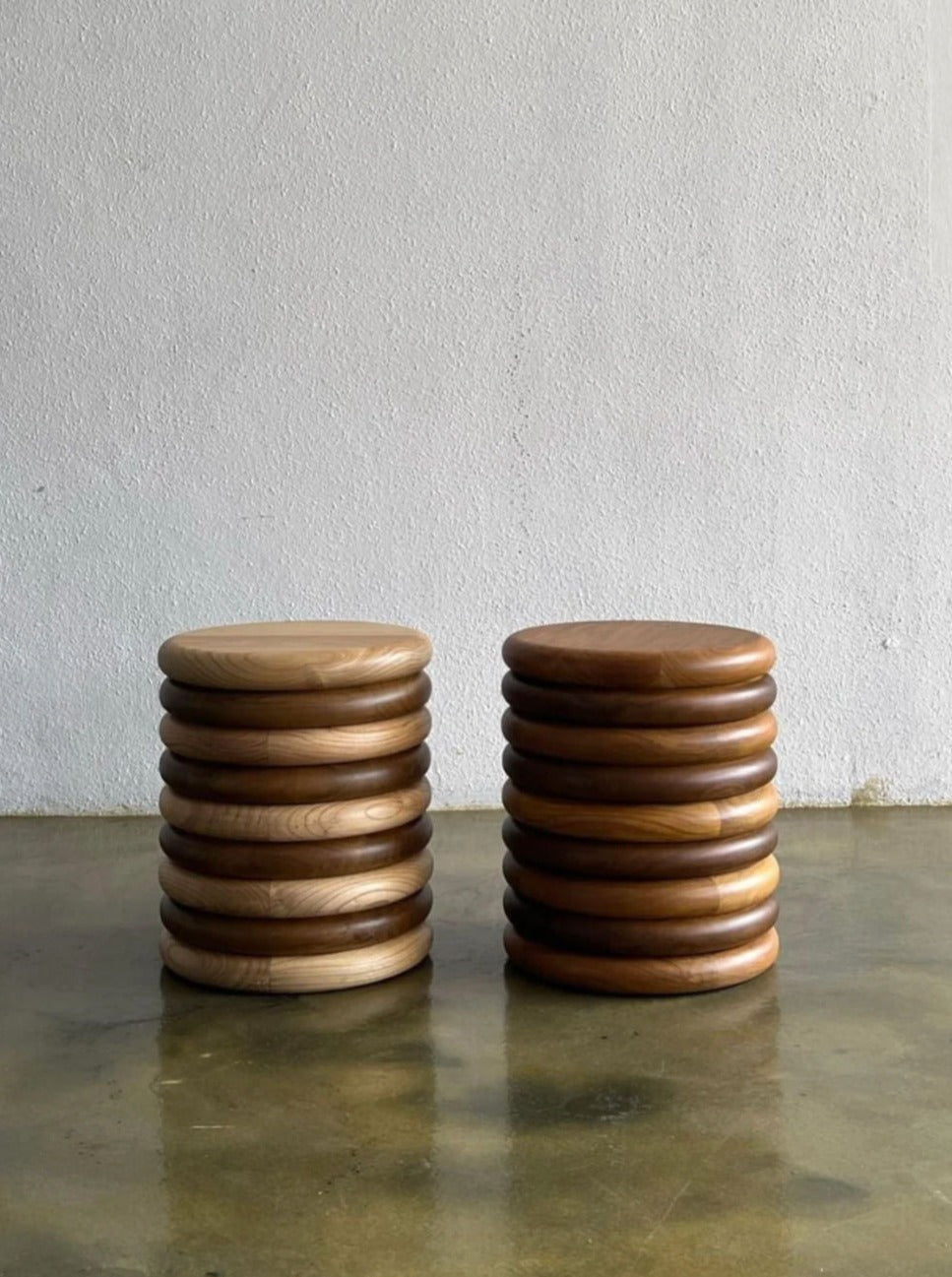A minimalist image showing two stacks of handcrafted wooden Hilu Stools / Side Tables by Studio Kallang, each stack consisting of 10 stools. The stack on the left, made from solid teak wood, is lighter, while the one on the right is darker. The stools double as side tables and are placed against a plain white wall on a polished concrete floor.