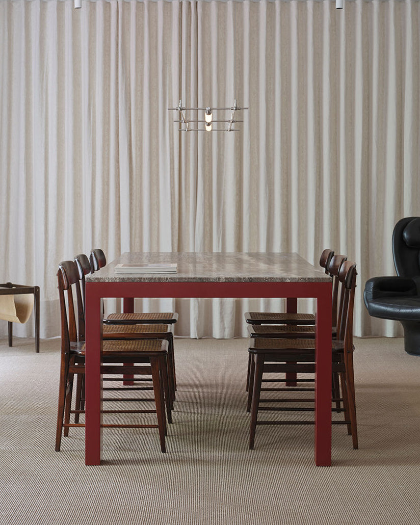 A modern dining room features the sleek Firme Dining Table from FEBRERO SHOP, with a dark red lacquered wooden frame and a stone top. It is surrounded by eight wooden chairs with woven seats, complemented by a unique pendant light, beige curtains, and a black armchair on the right.
