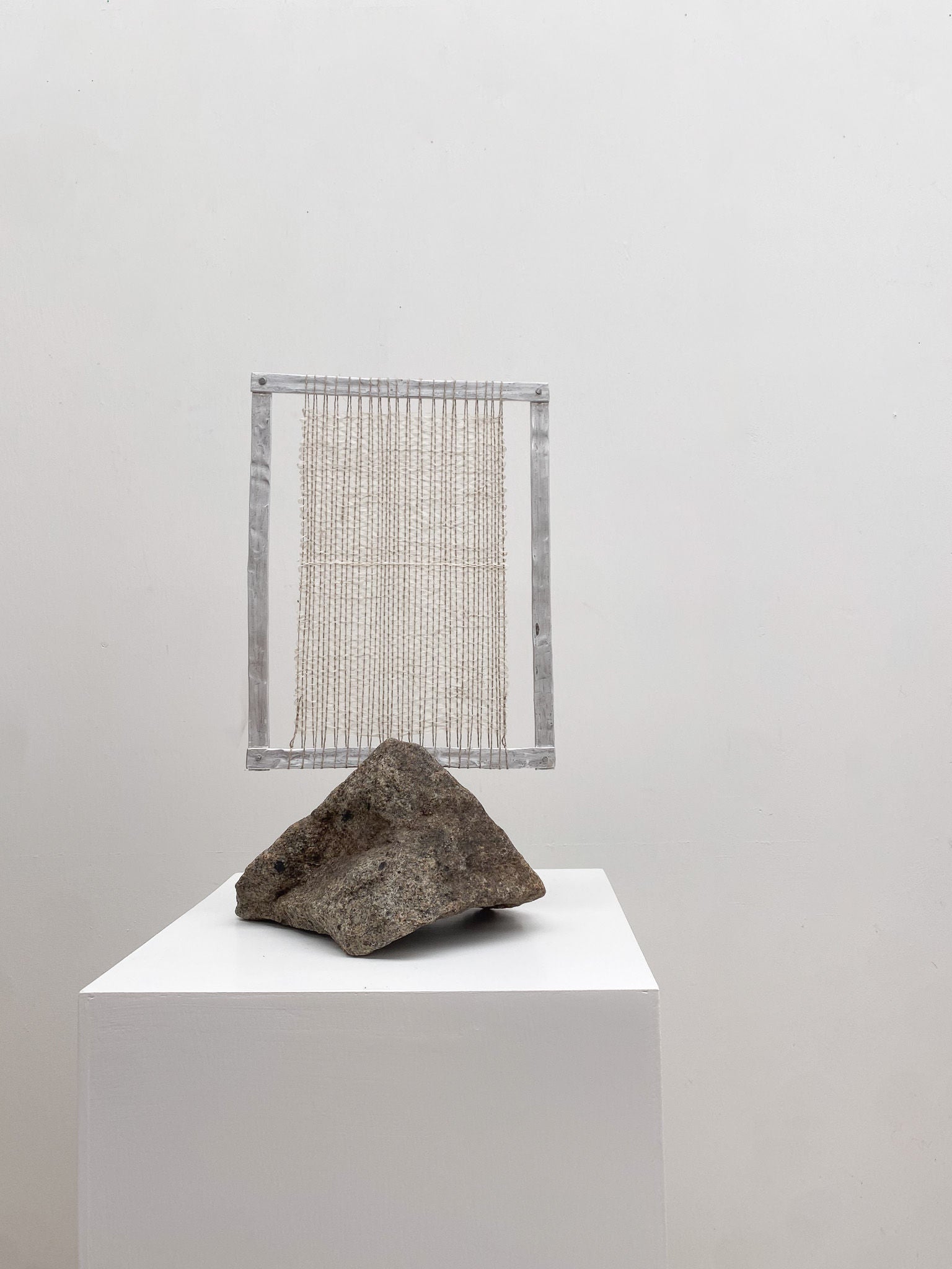 A piece of modern art featuring a textured granite rock atop a white pedestal, framed by a grid-patterned rectangular frame. The background wall reflects the serene glow of the Silver Moon I from Sundance Studio.