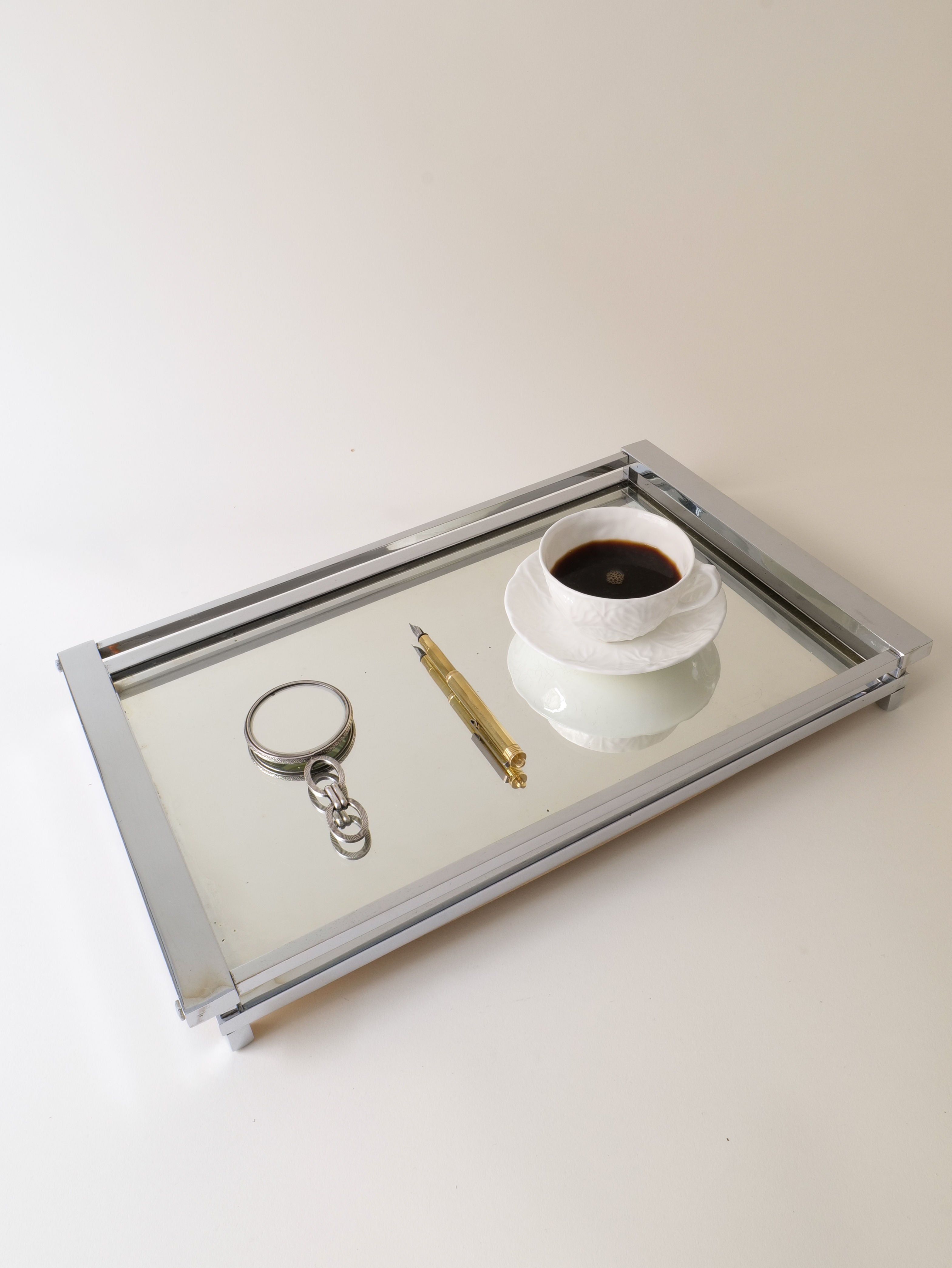 A Chrome Table Mirror from Collection apart echoes Bauhaus lines as it reflects a white cup of black coffee, a gold pen, and a silver magnifying glass with a chain. The simple elegance is reminiscent of French C1930-40s design against the plain white background.