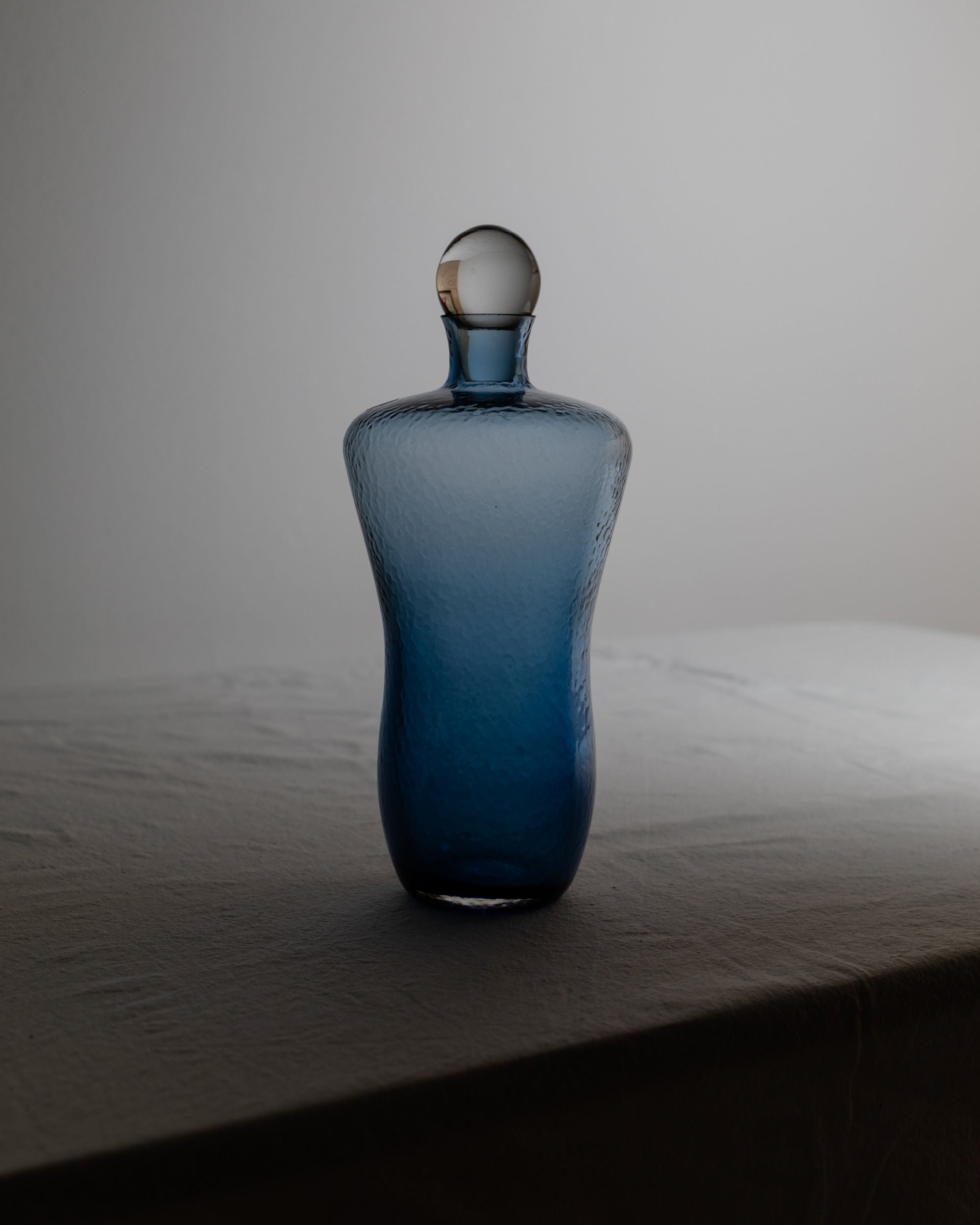 On a softly lit table, the Vintage Glass Decanter in Blue by Bottega Jacobs, featuring a round stopper, casts a subtle shadow on the fabric surface beneath it. The blue hue evokes a Hollywood Regency vibe against the plain light gray background.