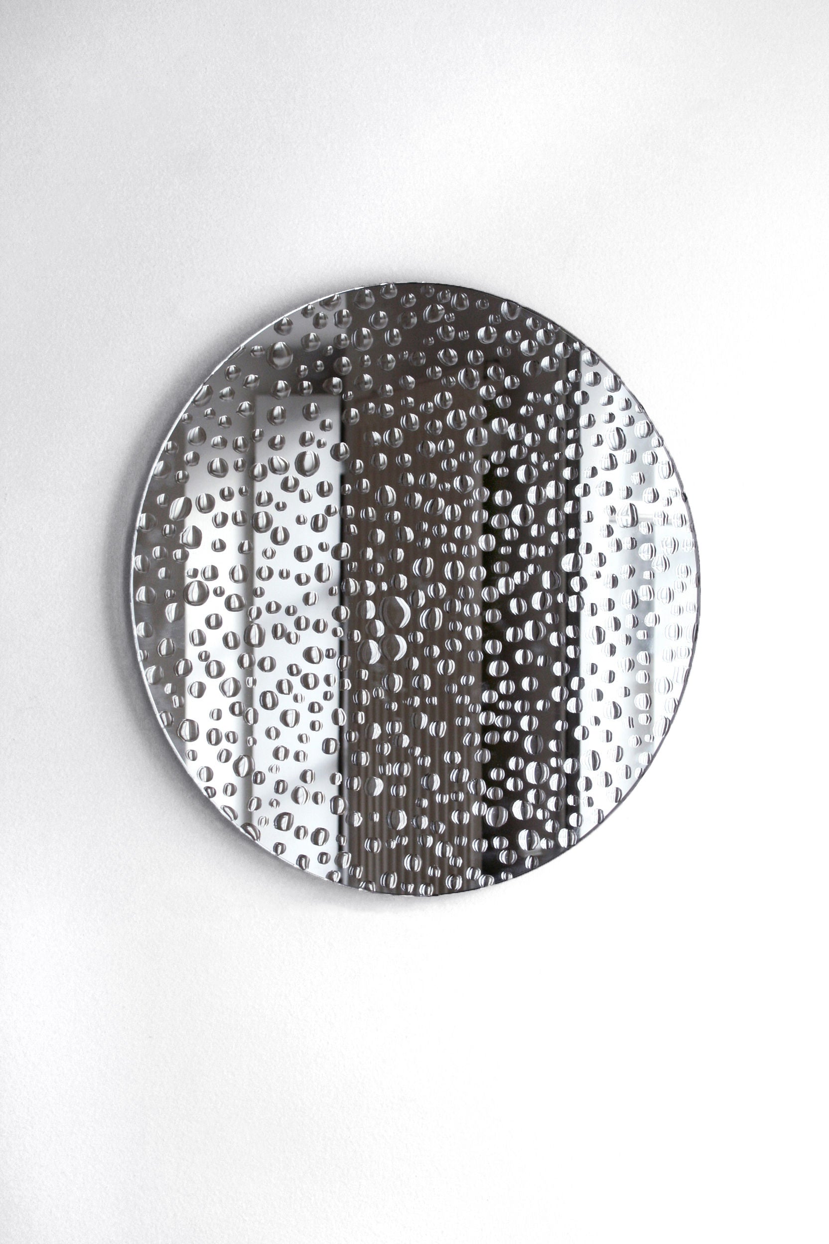 A round metallic mirror from Laurène Guarneri hangs on a white wall, showcasing an intricate pattern of raised circular bumps. This Mirror 11H20 features a water droplets design, creating a textured and reflective appearance that results in a unique visual effect. It truly stands out as an exceptional piece for any decor.