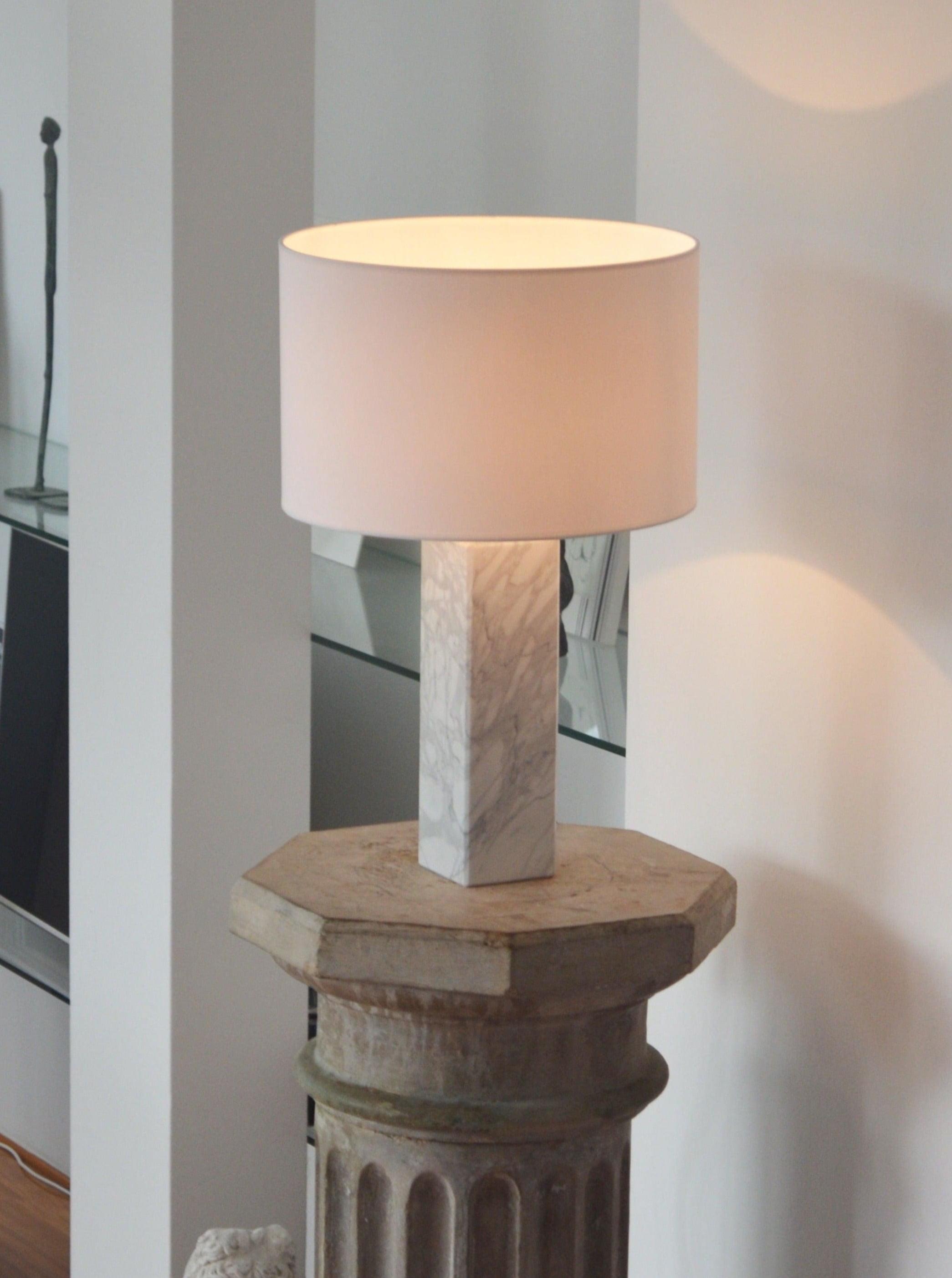 The Athens Table Lamp 1 by Brajak Vitberg shines forth with its white cylindrical shade and marble base, elegantly placed on a ribbed octagonal pedestal. It complements the luxury interior's soft lighting with its contemporary design.