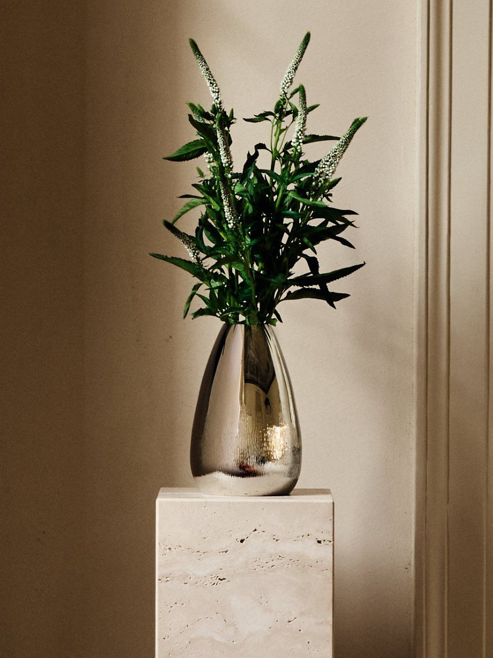 A zanetto Mexico Silver-Plated Vase, filled with green leafy stems and white flower spikes, makes a striking centerpiece on a beige marble pedestal against a pale wall.