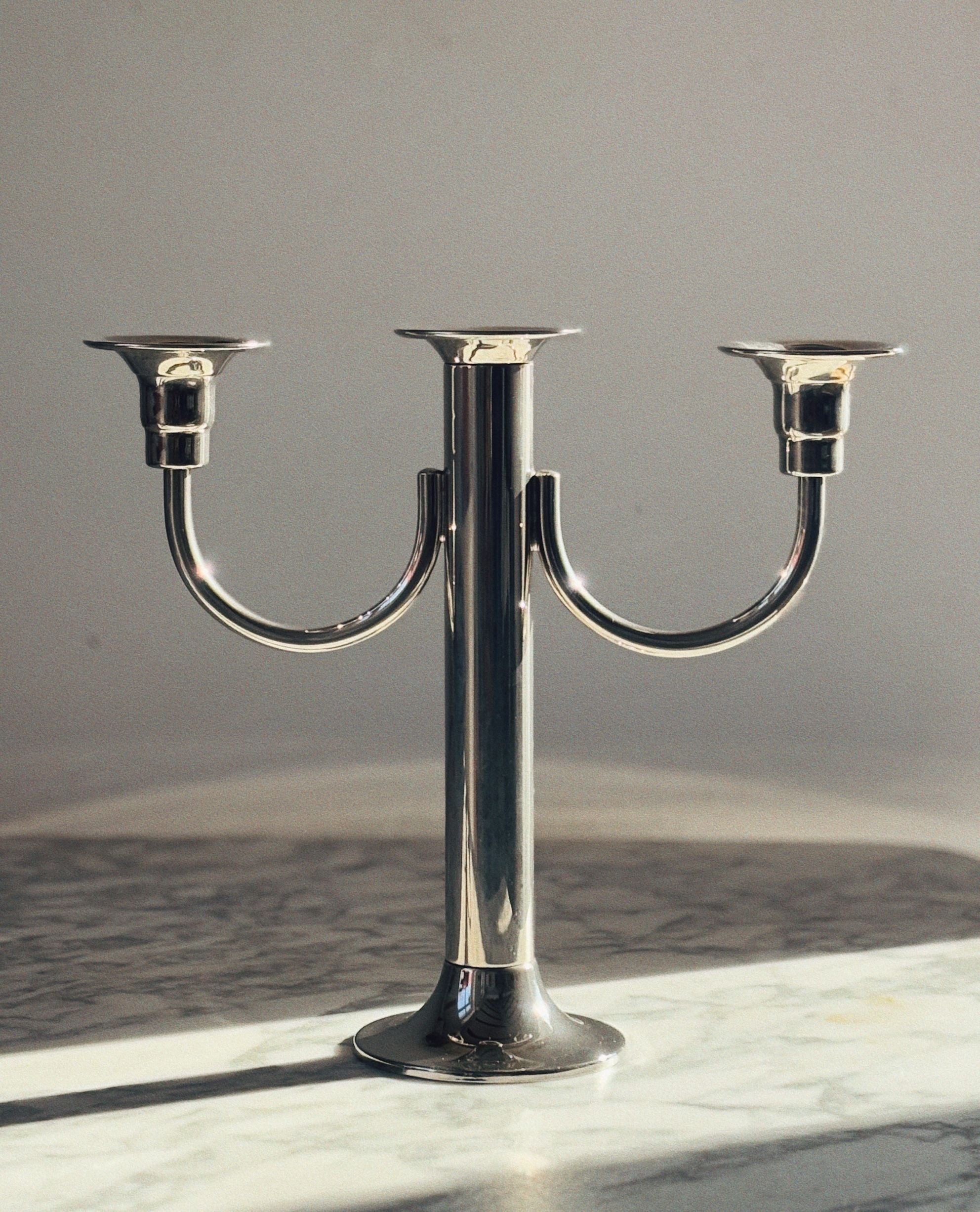 The Huerto 3 Arm Metal Candleholder, finished in silver, stands elegantly on a marble surface casting soft shadows against a plain neutral wall.