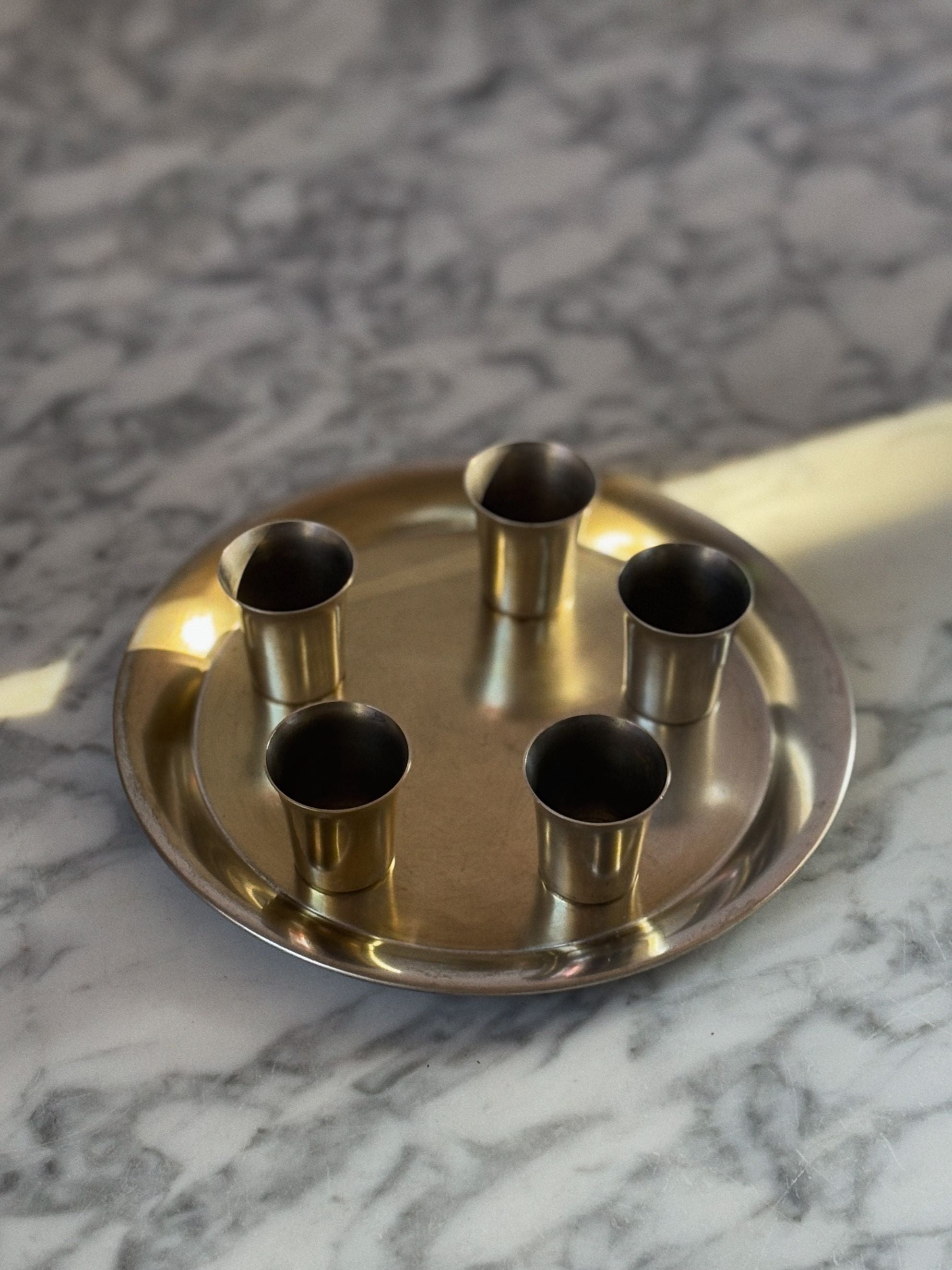 The Huerto Silver Plated Liquor Server Set includes a metal tray and five cylindrical liquor cups, all arranged on a marble surface. The round tray and cups reflect light, creating soft shadows that accentuate their artisanal craftsmanship.