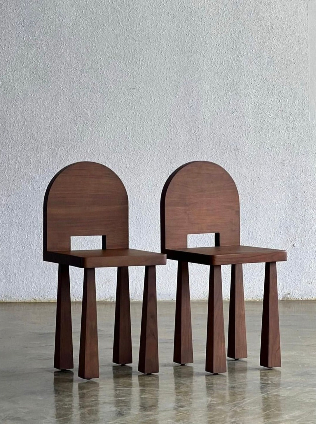 Two identical Luna Chair II by Studio Kallang with a modern design are placed side by side on a concrete floor. Each chair has a flat seat, a rectangular cutout in the backrest, and four flared legs that are angled outward. The background is a plain textured wall.