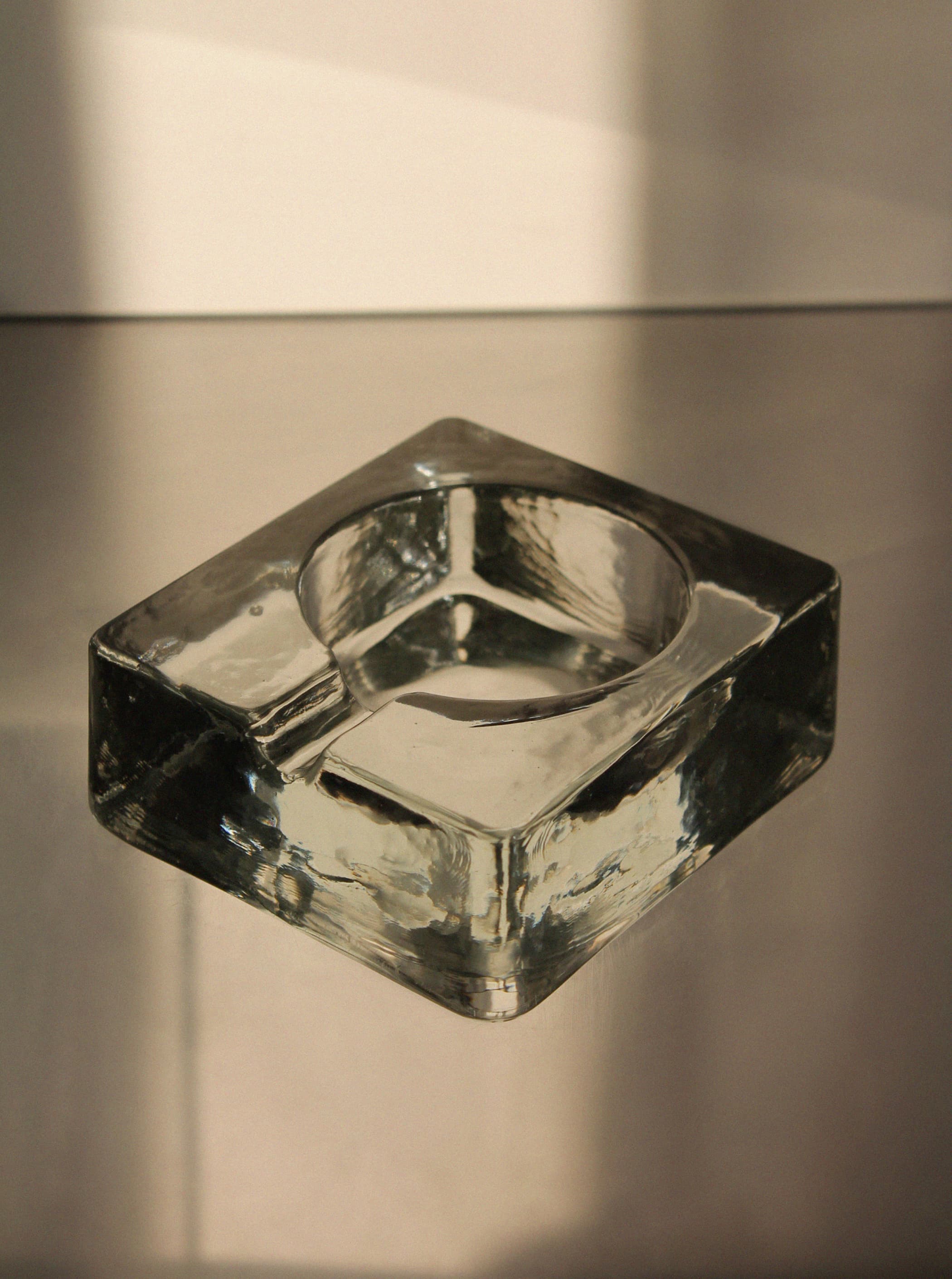 The Brutalist Massive Glass Ashtray Bowl by Vintage Stories, with its clear, square Scandinavian design, rests on a reflective metallic surface bathed in warm light, casting bold shadows and subtle highlights in true brutalist style.