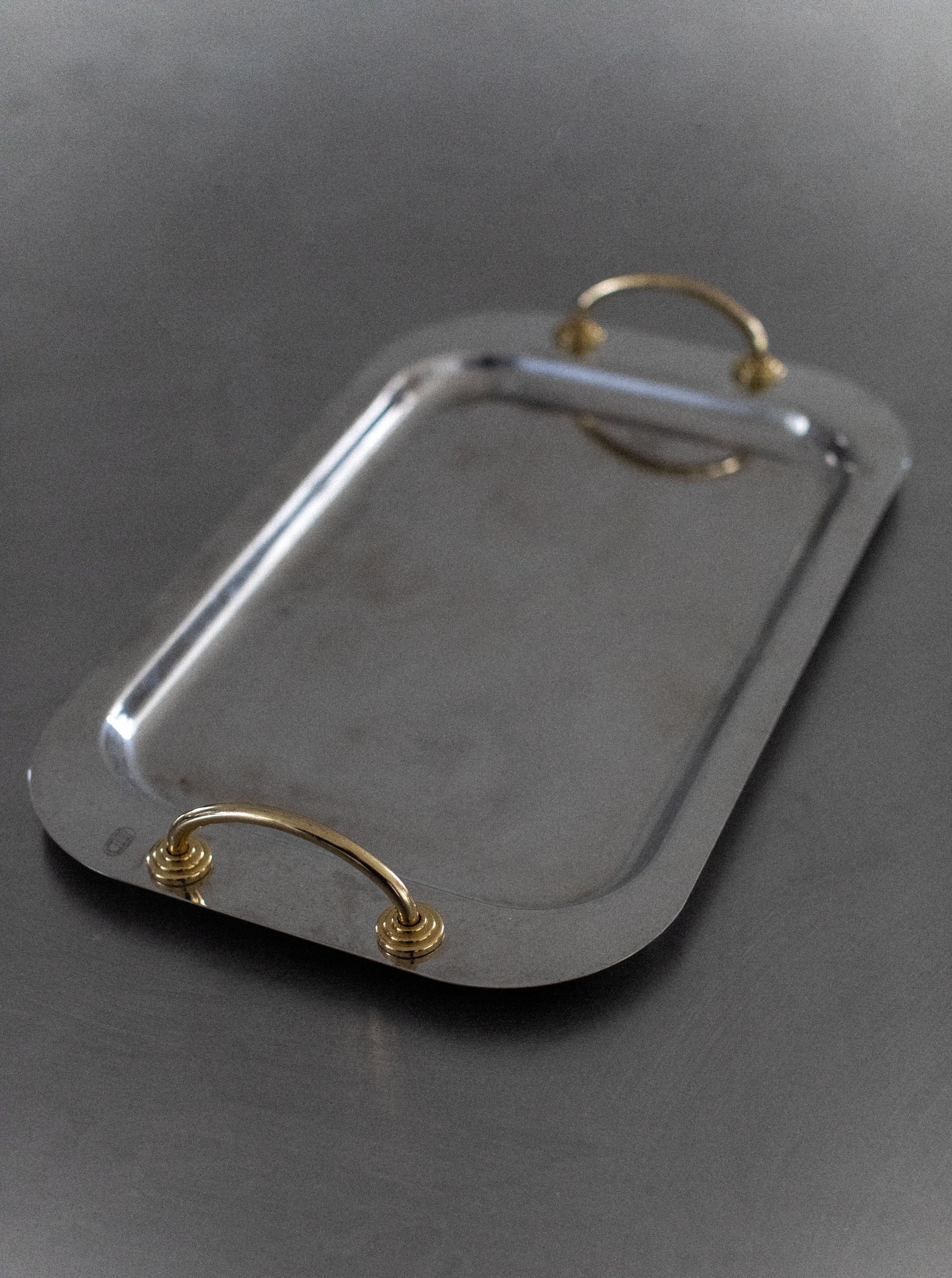 A rectangular Out For Lunch Stainless Steel Tray with rounded corners, featuring two gold-colored handles on either side. The tray rests on a grey surface, reflecting light faintly on its polished finish, showcasing its modern elegance and smooth, contemporary style.