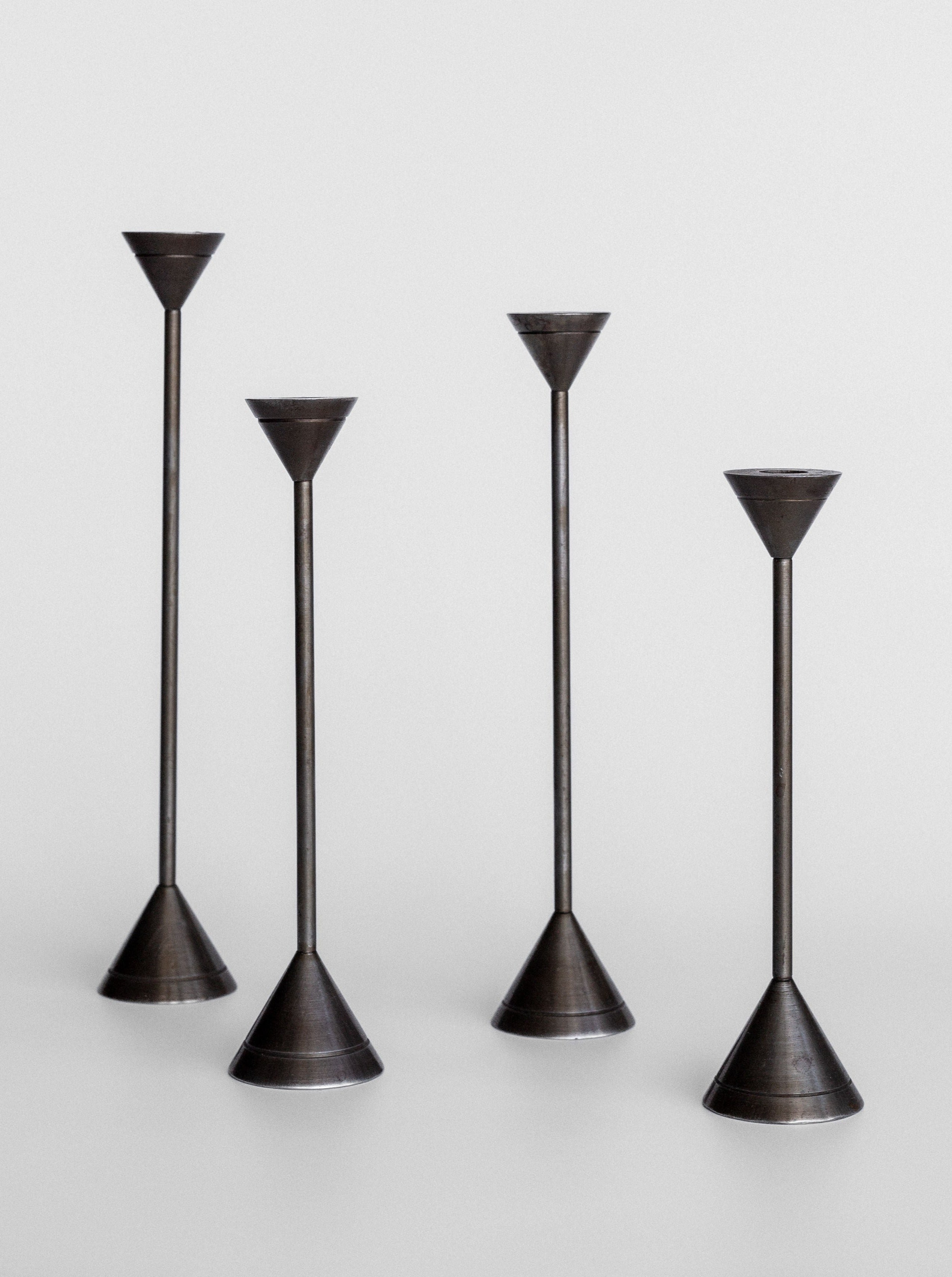 The Spigel Set of Four Postmodern Candle Holders are showcased against a light grey background. Each candle holder features a minimalist, solid metal construction with varying heights, conical bases, and slender stems leading to small, funnel-shaped tops designed to hold candles. Their sleek design exudes postmodern aesthetics.