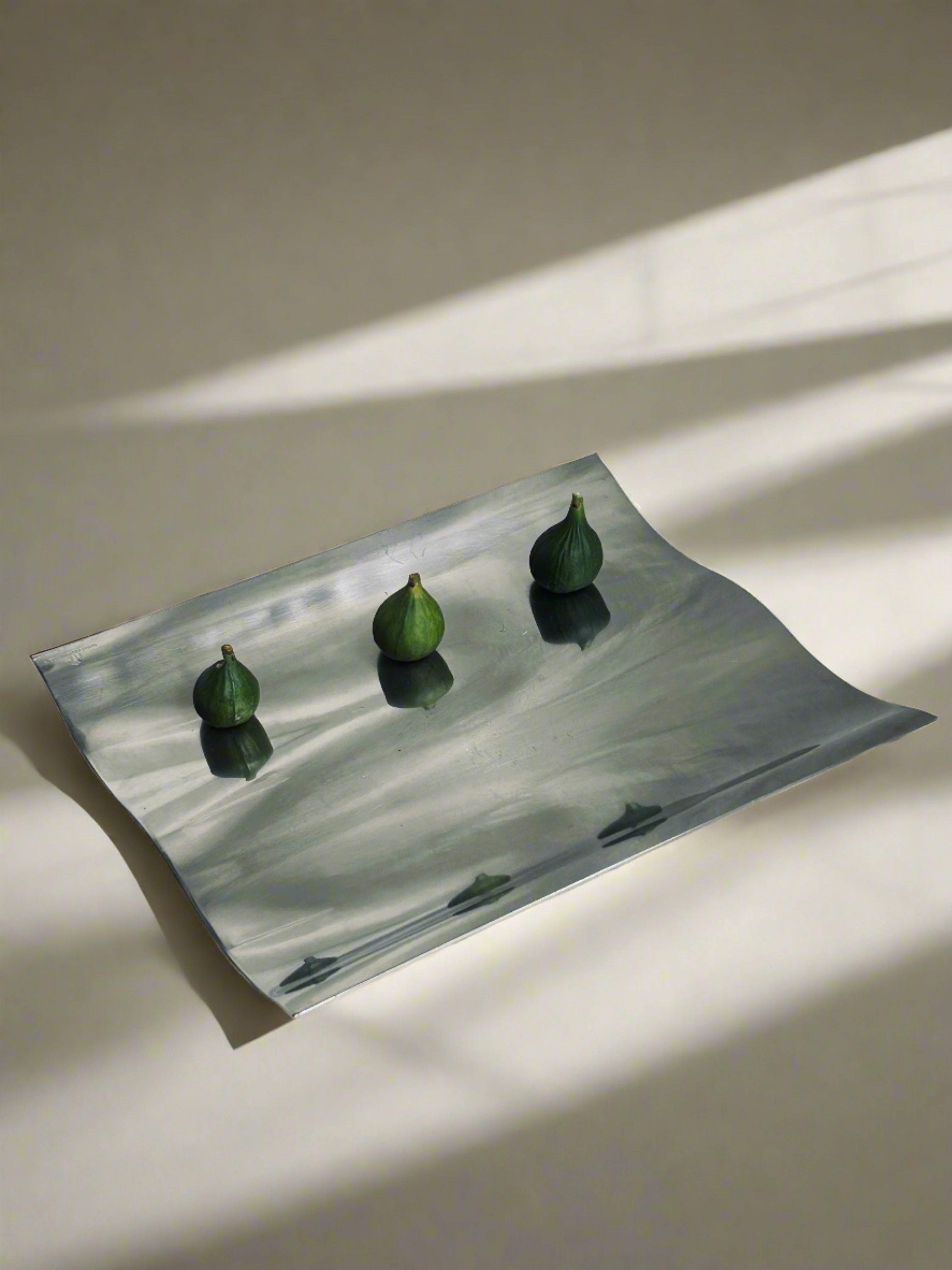 The elegant Wave Tray by Royal Copenhagen from Flock Studio, crafted in stainless steel, holds four small green figs and casts subtle shadows on a neutral-toned surface. Soft light and shadows form an elegant, minimalist composition with a modern twist.