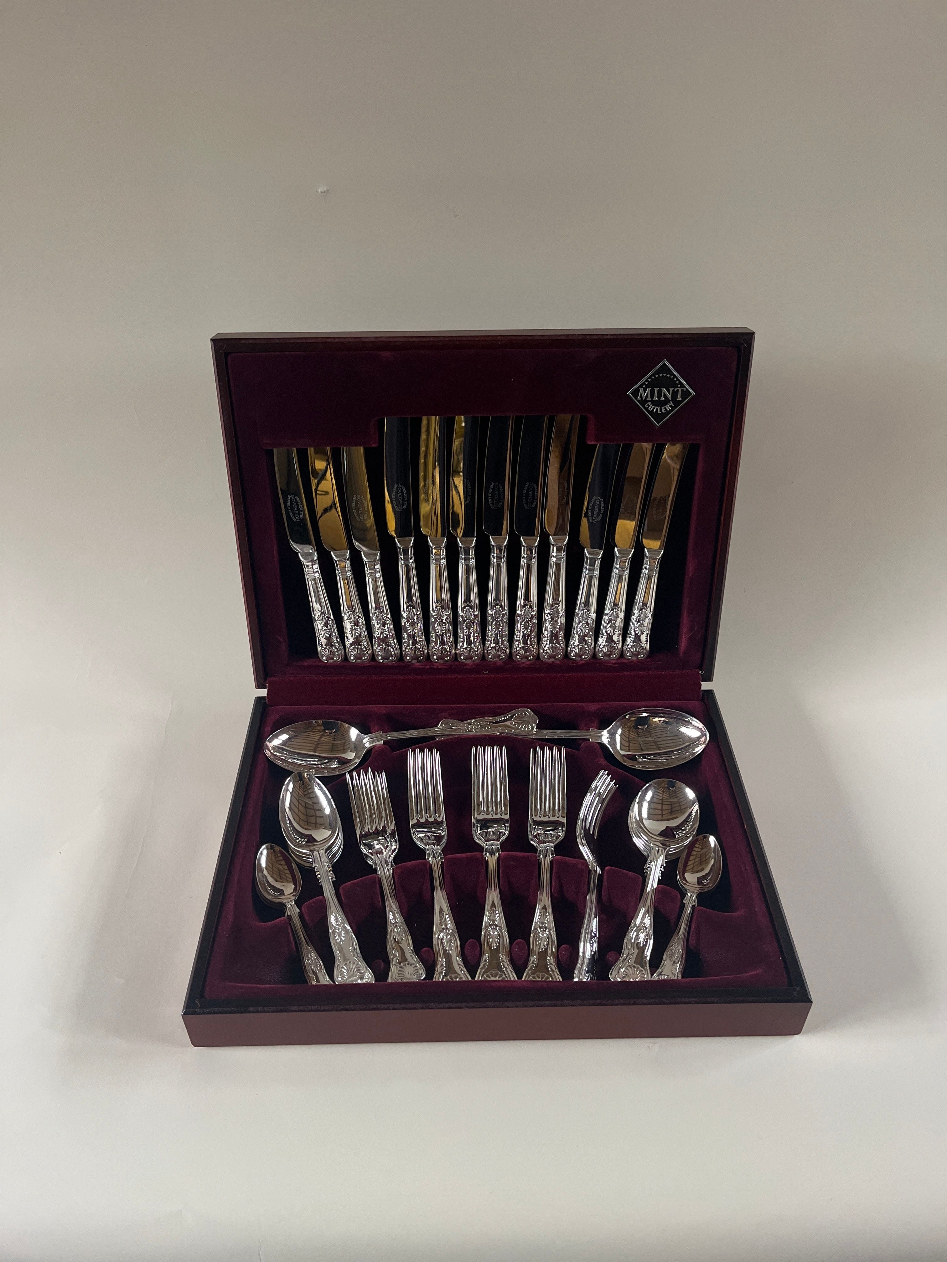 The Maud Vaughan 44 Piece Boxed Silver Service with Shell Motif offers a collection of silver-plated spoons, forks, and knives, characterized by their intricate handles. Encased in luxurious red velvet, this boxed flatware set boasts a prominently displayed logo on the upper right corner of the lid.
