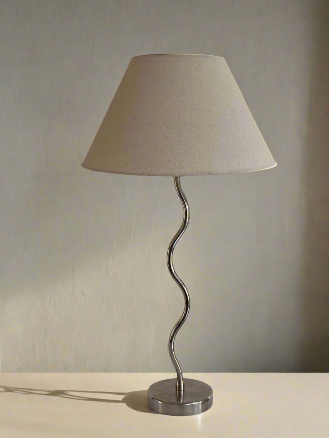 The Maud Lamp - Nickel by Maud Vaughan features a modern beige conical lampshade atop a wavy nickel-plated stand, set on a light surface against a neutral wall, showcasing exceptional craftsmanship.