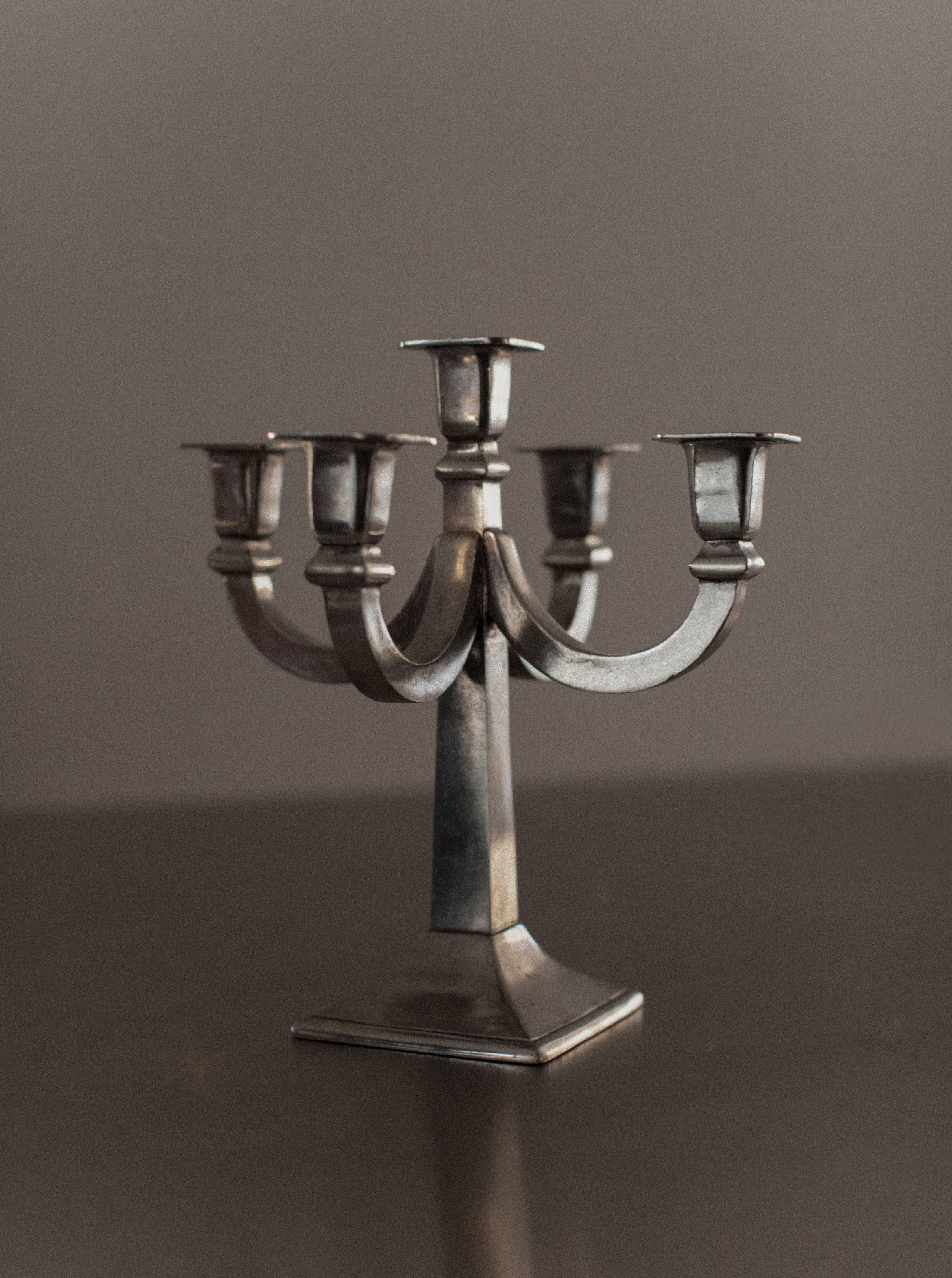 A vintage metal candelabrum with five candle holders arranged in a symmetrical design. Standing on a polished flat surface with a plain dark background, it boasts a slightly tarnished appearance, adding to its distinctive charm. This Mid-century Candleholder by Out For Lunch makes an elegant addition to any home decor.