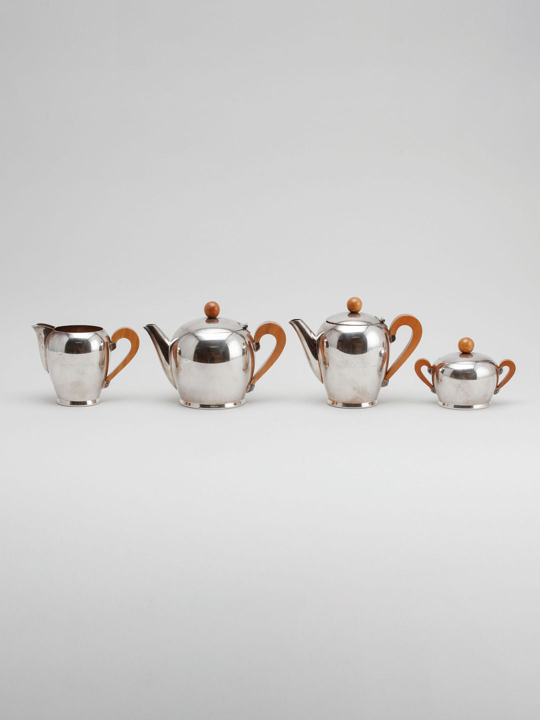 A clean, minimalist image of the Art Deco Tea/Coffee Set ‘Bombè’ by Alessi, available at Dodo Vintage, showcases mid-20th-century craftsmanship with its stainless steel finish and wooden handles and knobs. From left to right, it includes a creamer, teapot, another teapot, and a sugar bowl, all arranged in a line against a light grey background.