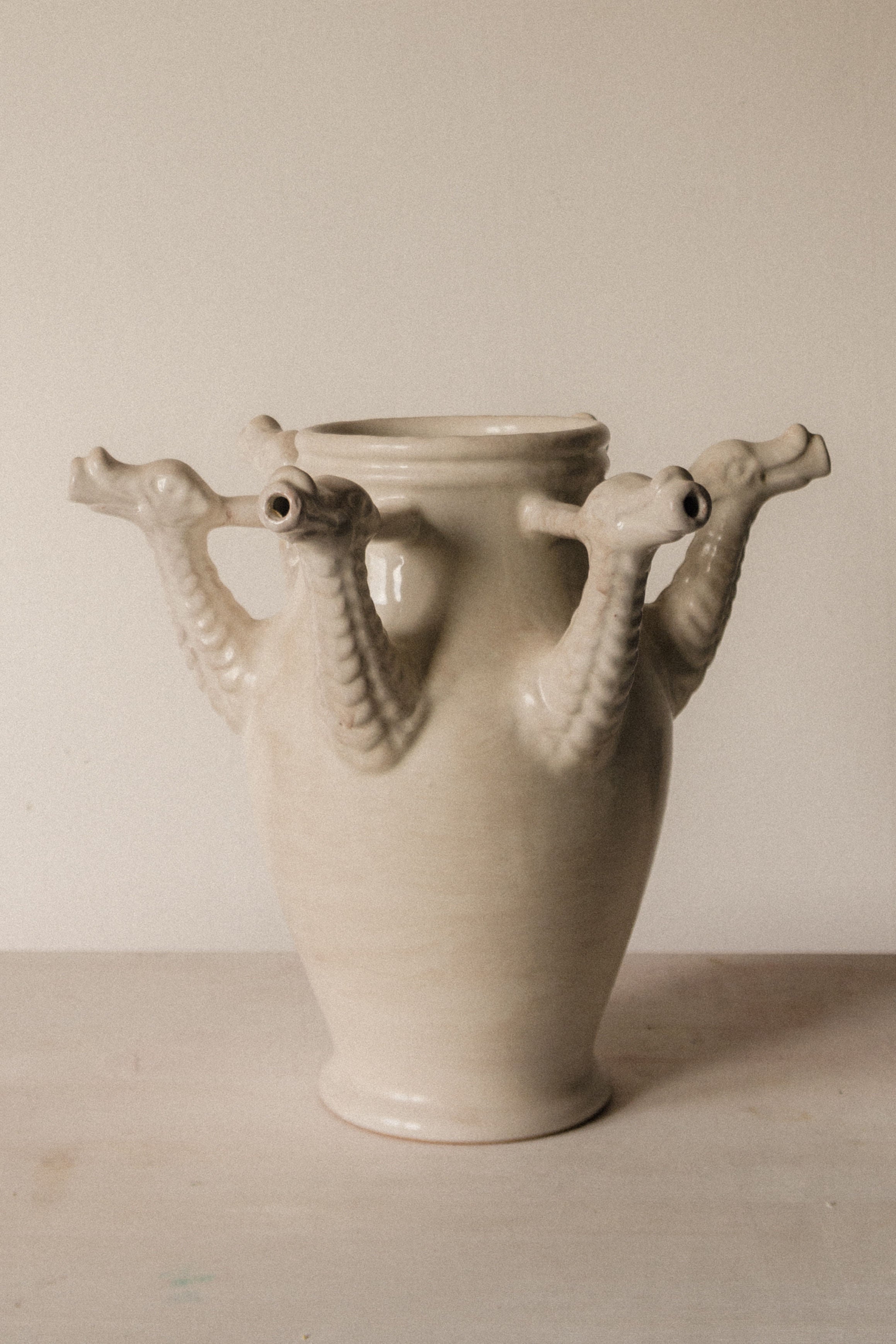 The Drago Bianco Vase by COSEINCORSO is a handmade beige ceramic piece from Italy, inspired by Castelli designs, featuring four outward-facing sculpted dragon heads near the rim and displayed on a plain surface with a neutral background.