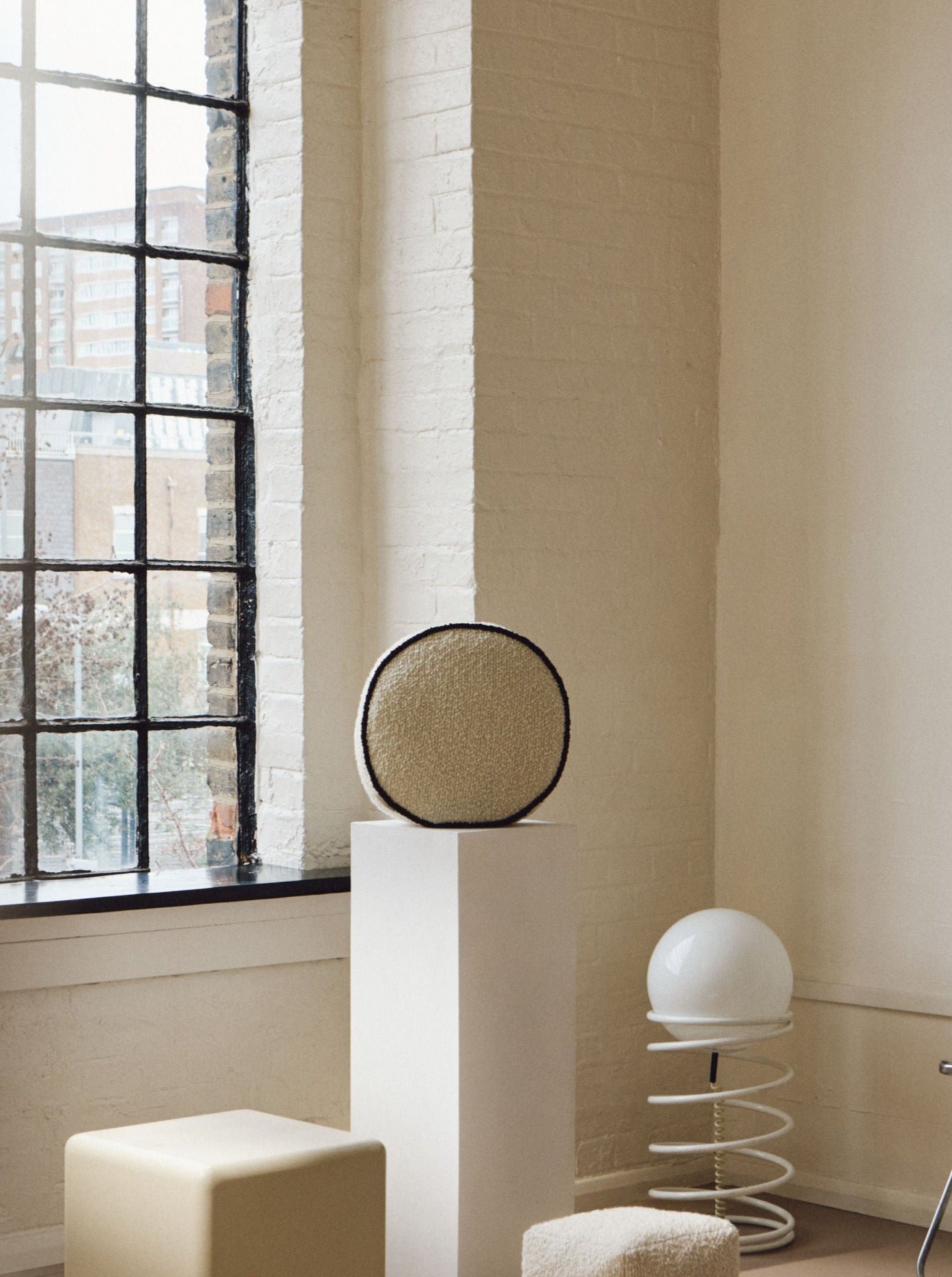 A minimalist room with a large window, light-colored walls, and assorted modern furniture. There's an AMÉLIORÉ Le Rond - Passepoil - Wool Bouclé - Egg White/Chocolate pedestal with a circular cushion made from luxurious wool bouclé, a cube pouf, a smaller rectangular pouf, a circular lamp with a spiral base, and a chrome chair with a cushioned backrest.