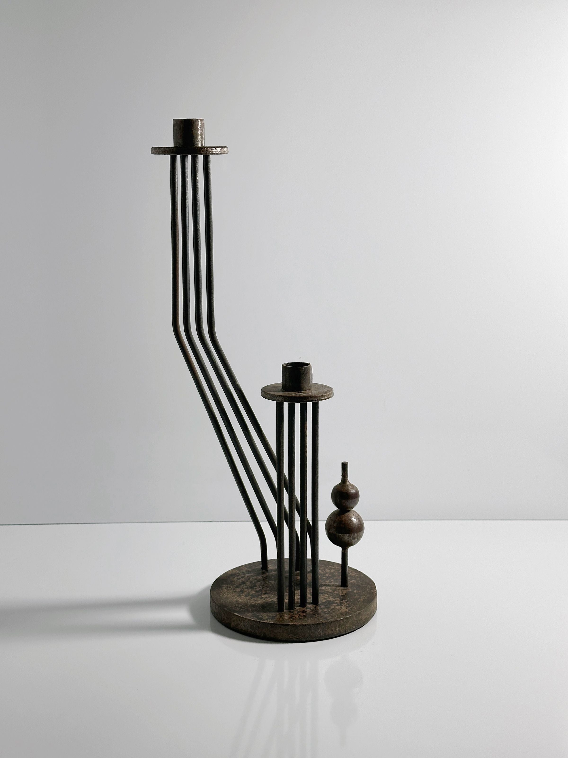 The Dodo Vintage Solid Brutalist Metal Candleholder from the 1960s stands on a white surface, featuring vertical rods, two circular platforms, and abstract shapes. Its rust patina and design cast intriguing shadows reminiscent of a minimalist sculpture.