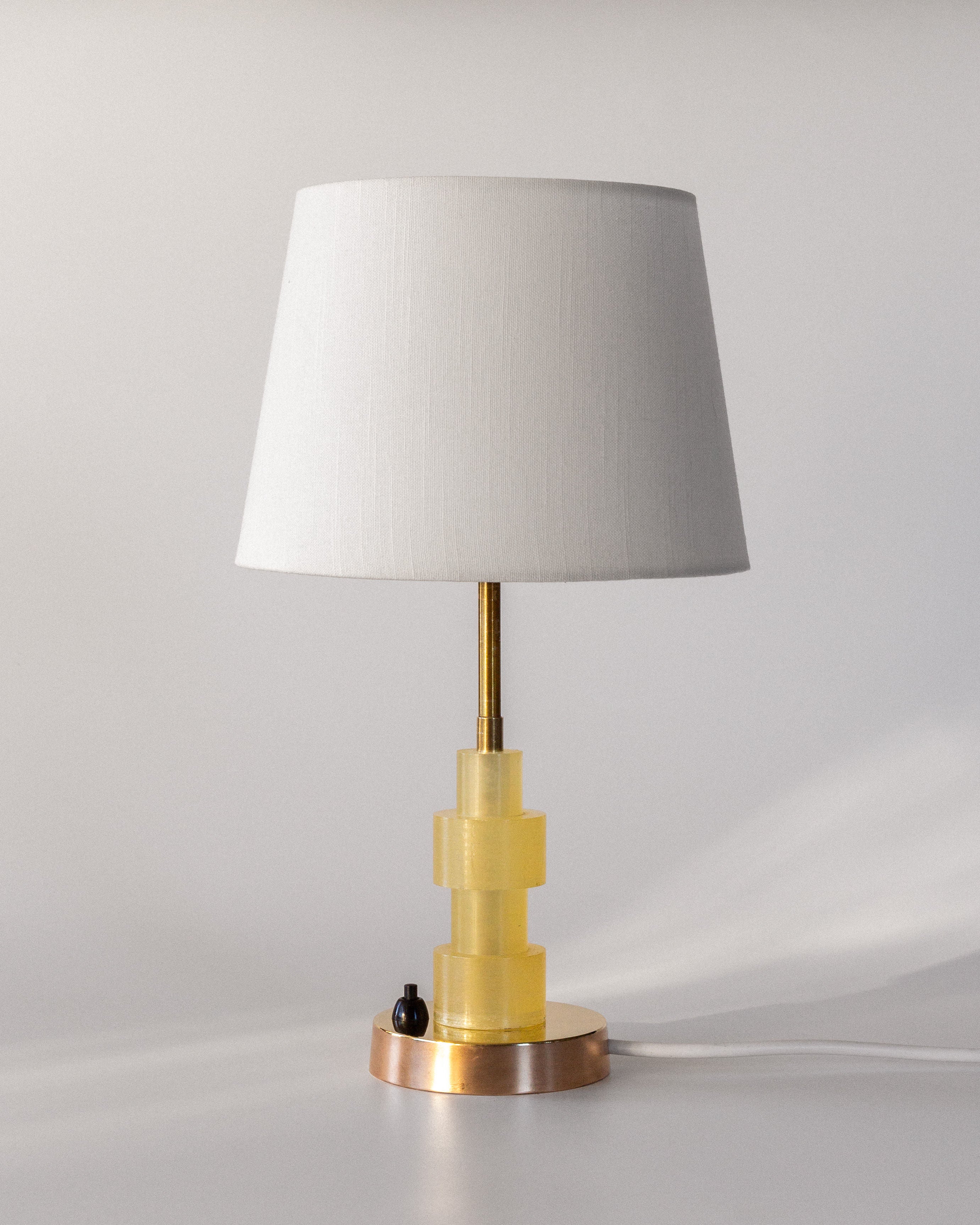 The Spigel Swedish Mid-Century Table Lamp features a white textile shade, a brass stand, and a geometric yellow base with copper-colored accents. It showcases exquisite Scandinavian design and includes a small black switch at the bottom.