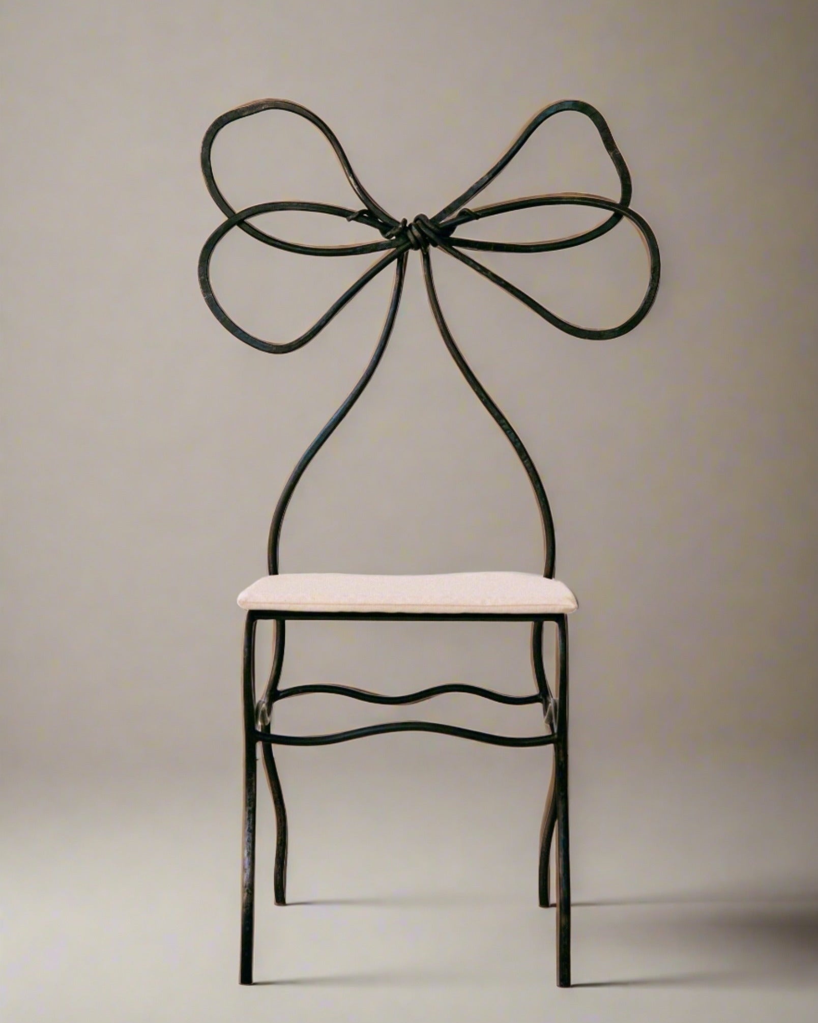 A minimalist chair with a unique, artistic backrest made of twisted iron forming a bow shape, The Bow Chair by Worn Studio. The seat is covered in a light, soft material, contrasting with the stark, metallic frame. Perfect for collectors and design amateurs alike, this high-end design piece stands against a plain, neutral background.