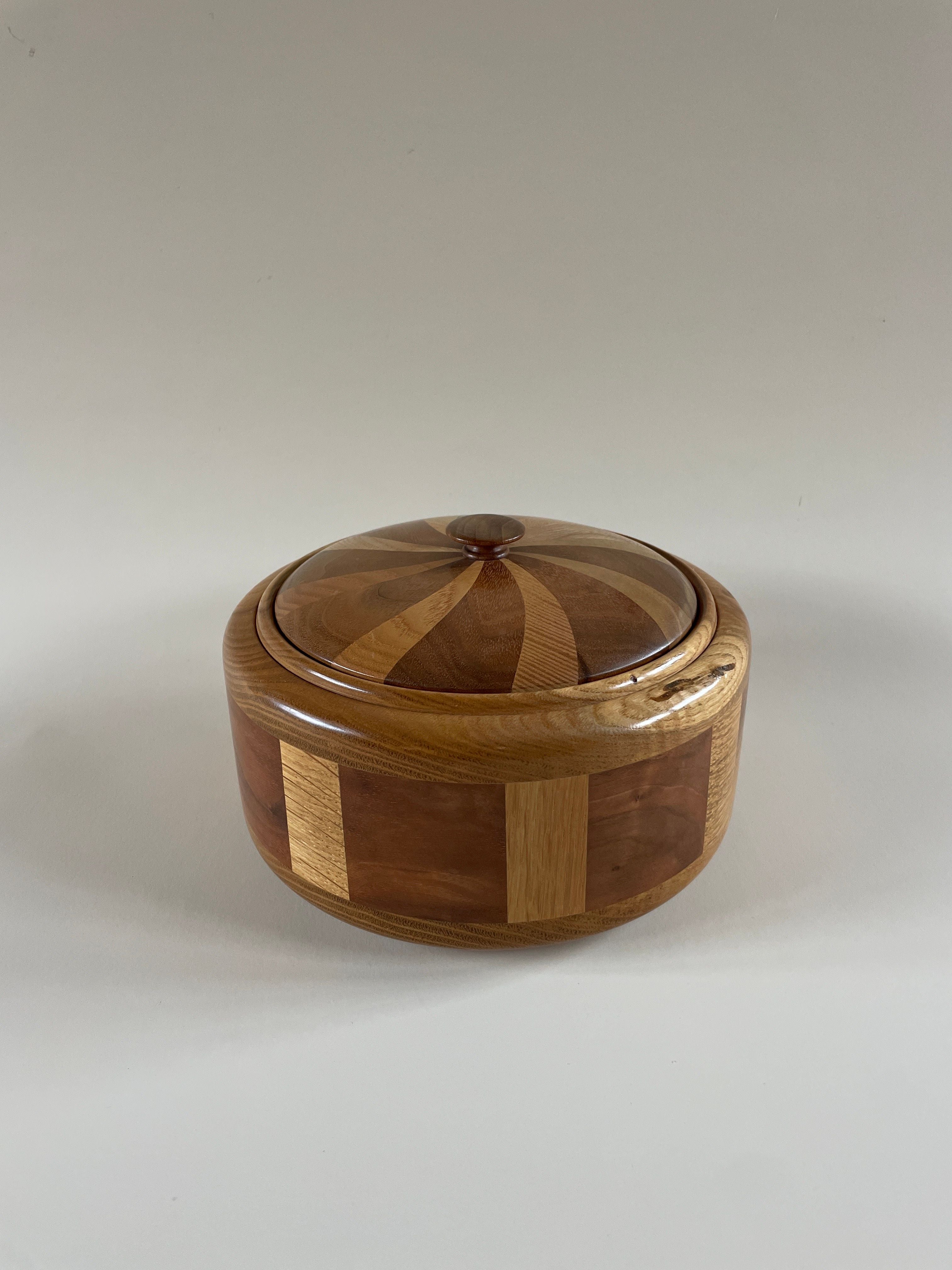 The Hand Turned Lidded Circular Box by Maud Vaughan is a skillfully crafted wooden box featuring a lid with an intricate geometric pattern in alternating light and dark wood tones. The design on the lid creates a striking striped effect, while the sides showcase a varied panel pattern, all set against a plain white background.