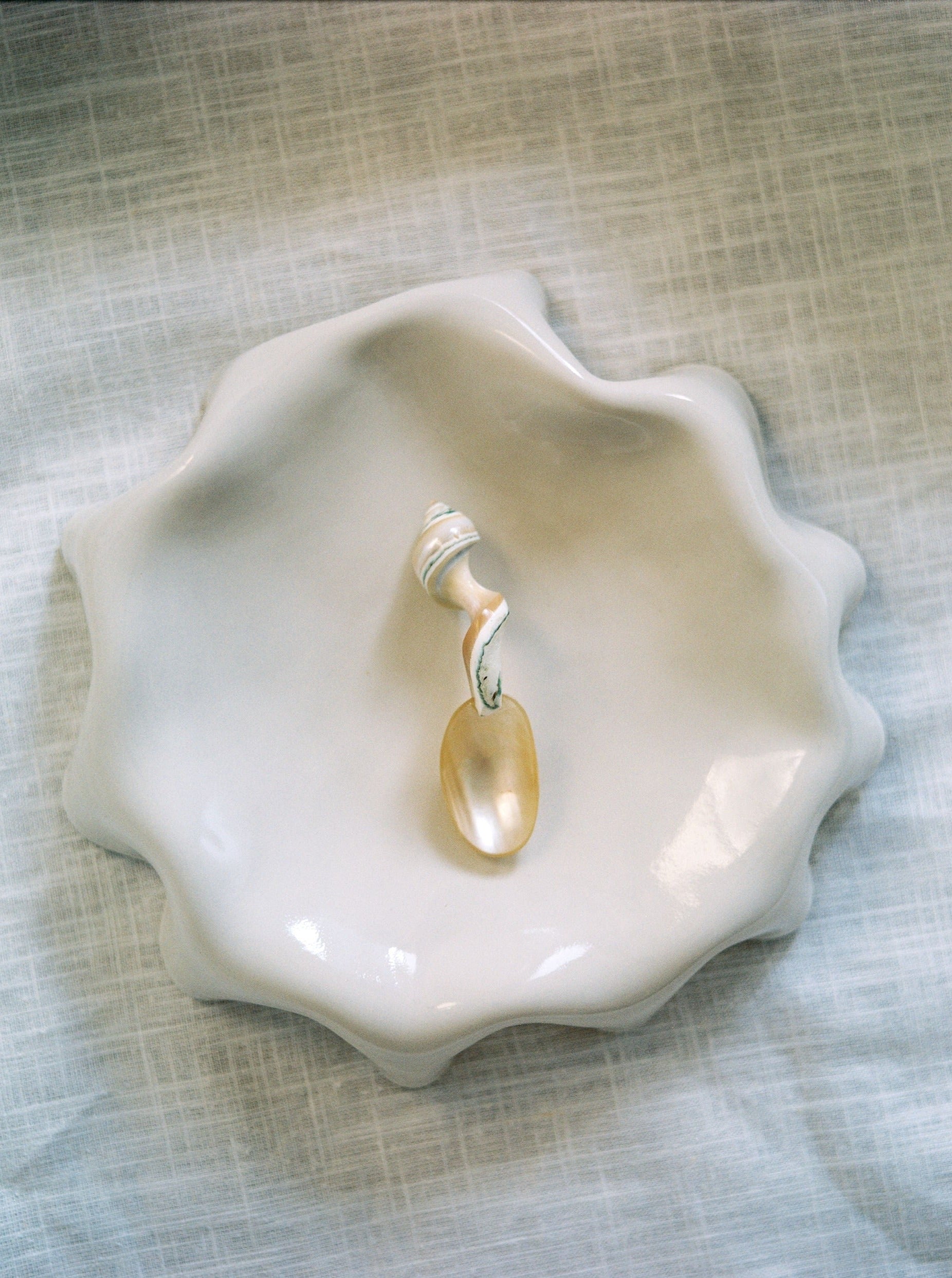 A wavy-edged, glossy Melted Butter Plate in Off White from LESS graces a light, textured surface. Resting within the dish is a small spoon with a handle crafted from a white seashell, displaying hints of beige and gray. This delicate coastal-themed aesthetic exemplifies practical tableware at its finest.
