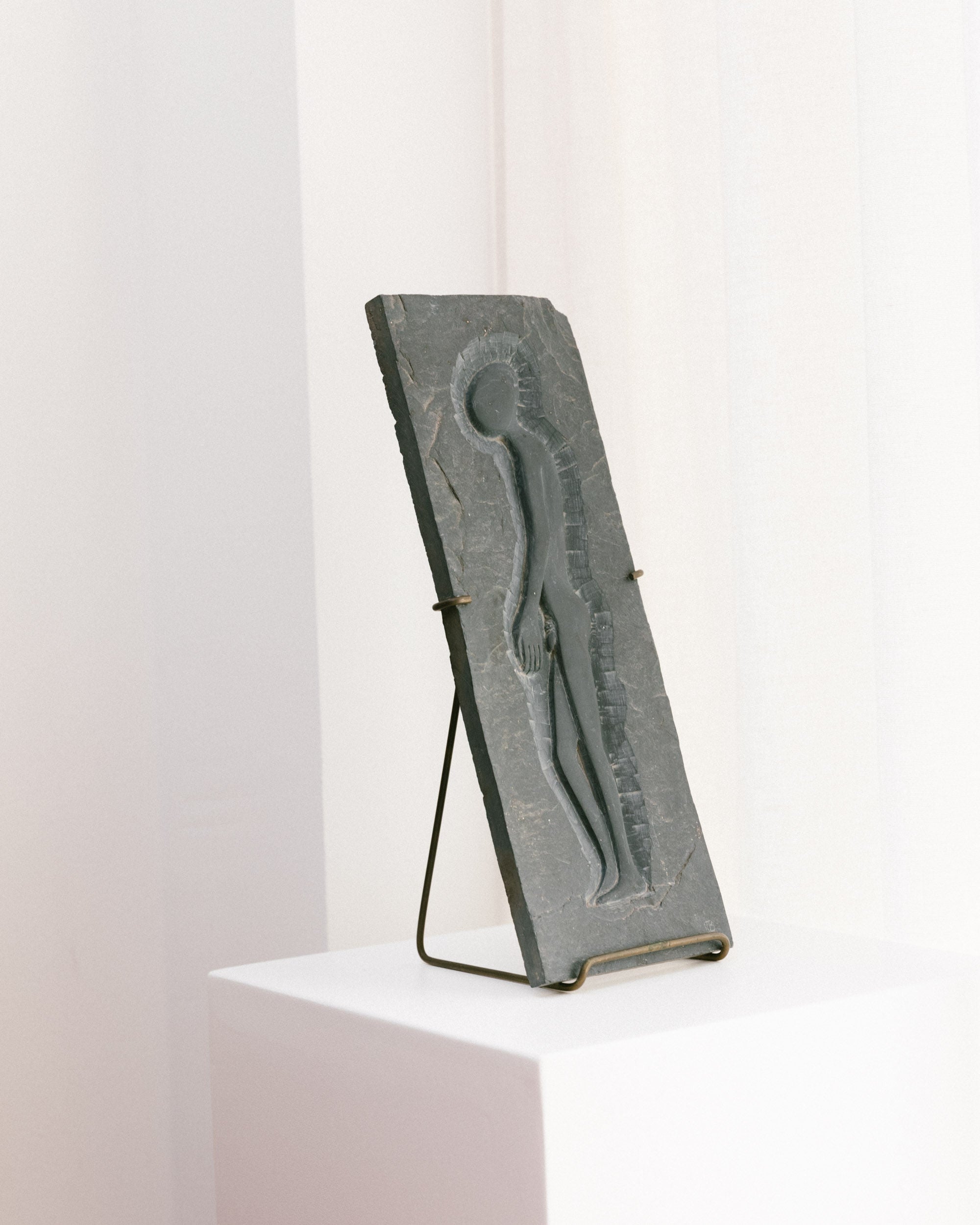 The Slate Art Panel - Alien Man 1970 by Bottega Jacobs features a minimalist human figure in relief carved into a rectangular stone slab. Displayed on a stand atop a white pedestal, it is set against a soft, white background.