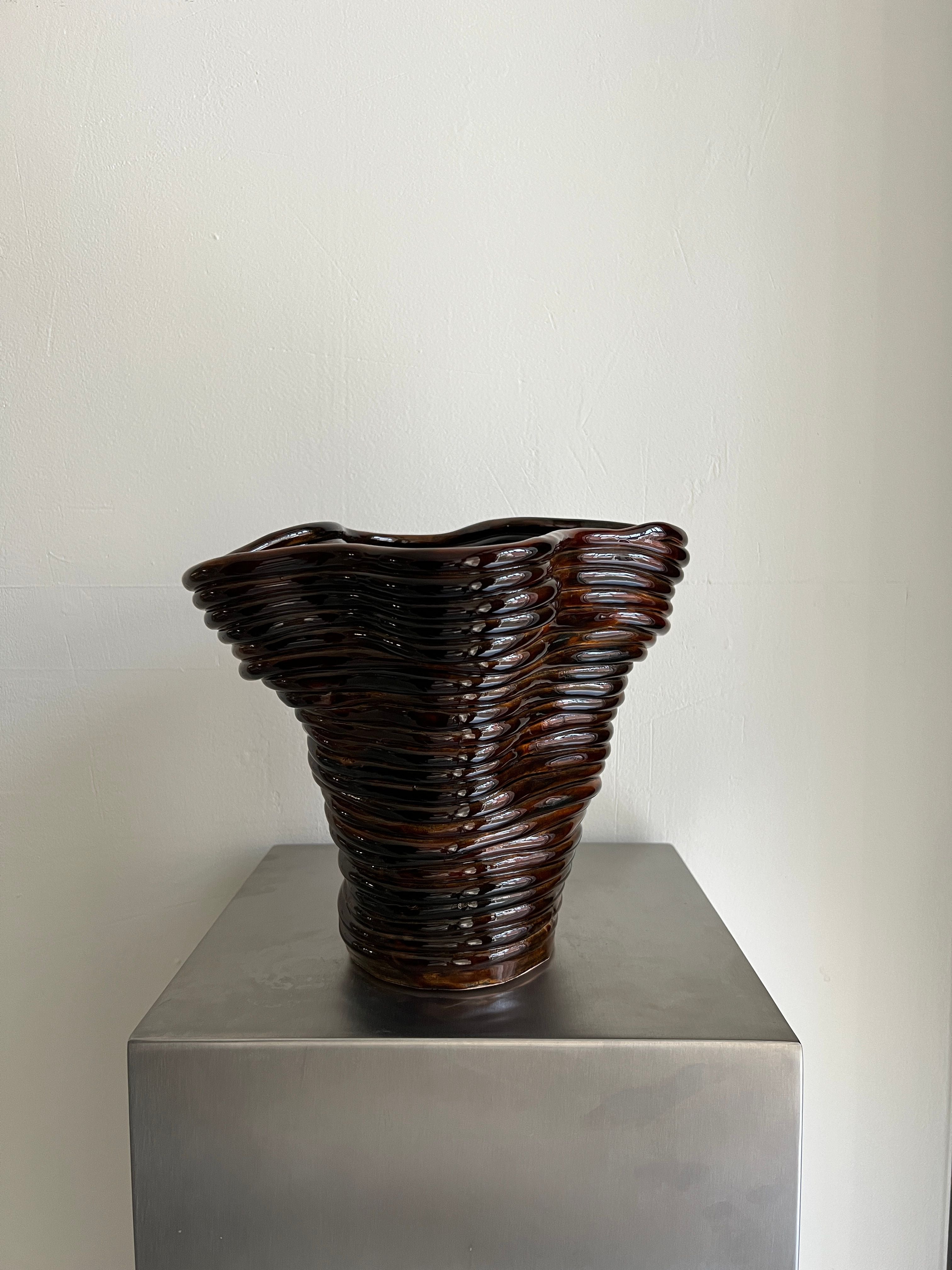 The Wobbly Vase Brown by 2222Studio, a handcrafted ceramic vase featuring a wavy ridged texture and dark brown flared design, stands on a metallic pedestal against a white wall, creating a stylish focal point.