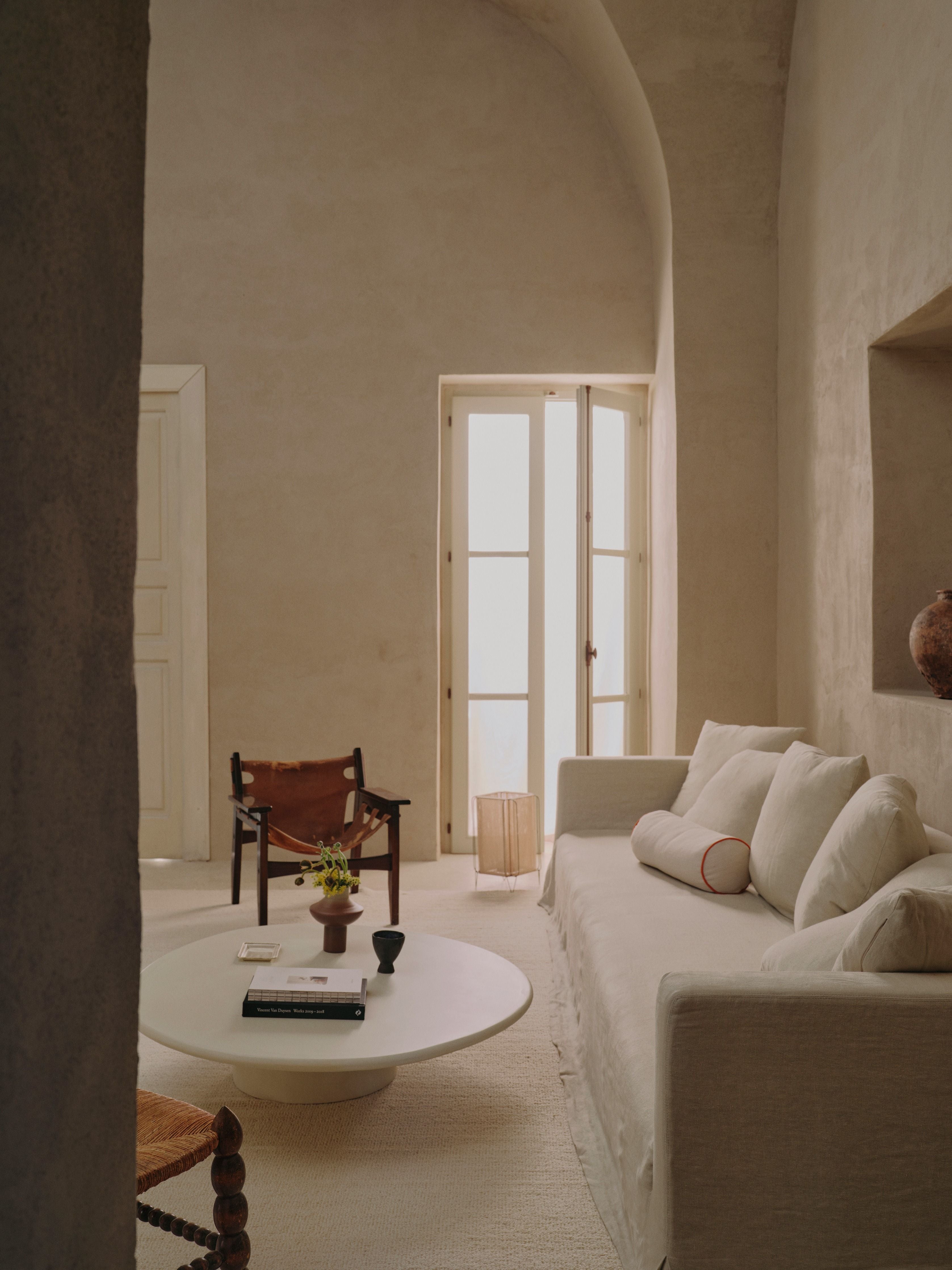 In a minimalist living room with neutral tones, a light beige sofa pairs with the MarlotBaus Cinzia Coffee Table. A brown leather chair with an organic silhouette completes the scene. Sunlight filters through sheer curtains onto a micro cement floor, where a small plant thrives.