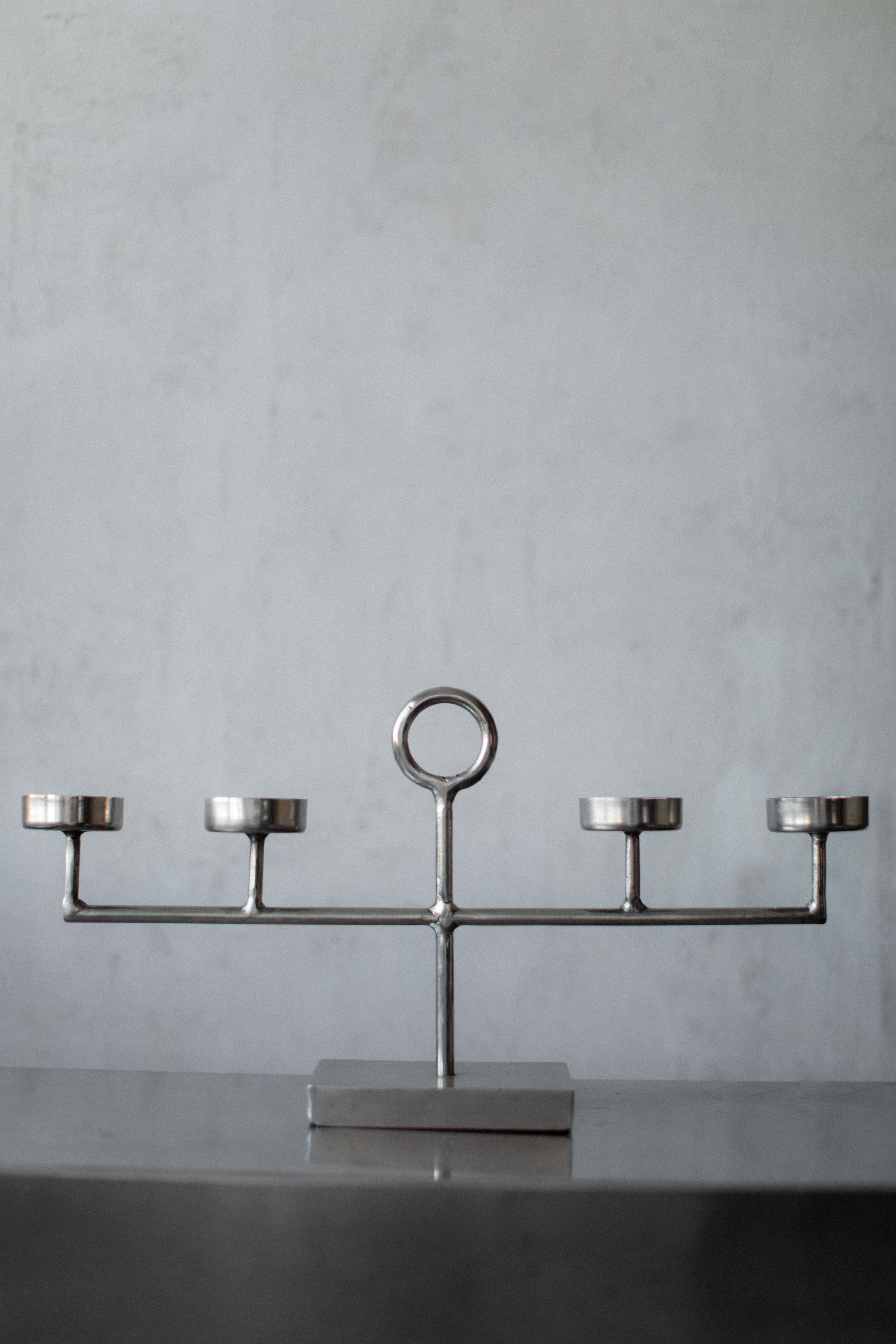 The Out For Lunch Steel Candleholder showcases a minimalist, geometric design with five symmetrically arranged holders and a central circular handle, elegantly resting on a smooth, dark surface against a light gray background.