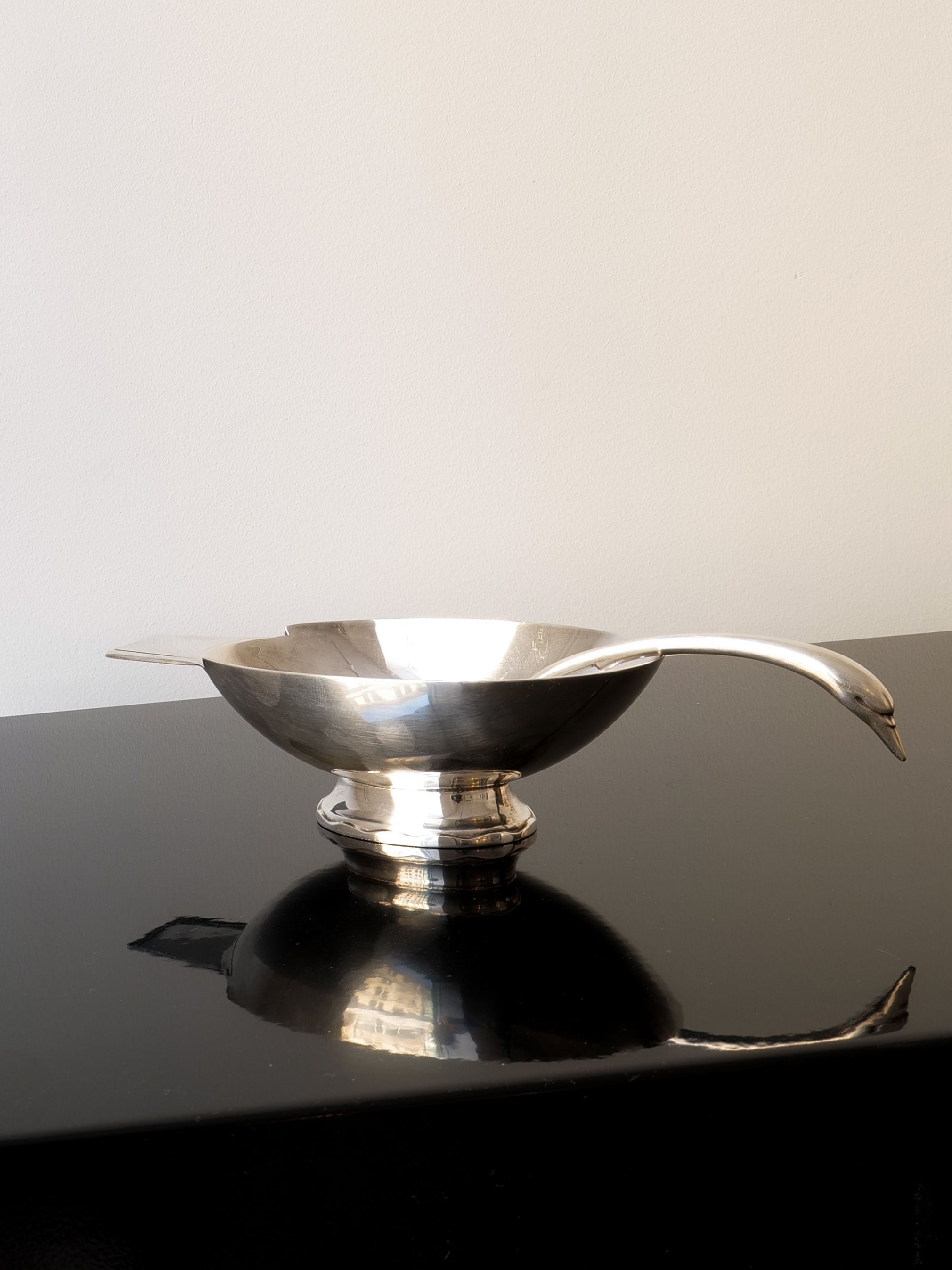 A gleaming Christofle Silver Swan Bowl & Spoon from Collection Apart rests on a glossy black surface, casting a soft reflection beneath it with its wide bowl and elegantly curved handle. The minimalist background is plain and light-colored.
