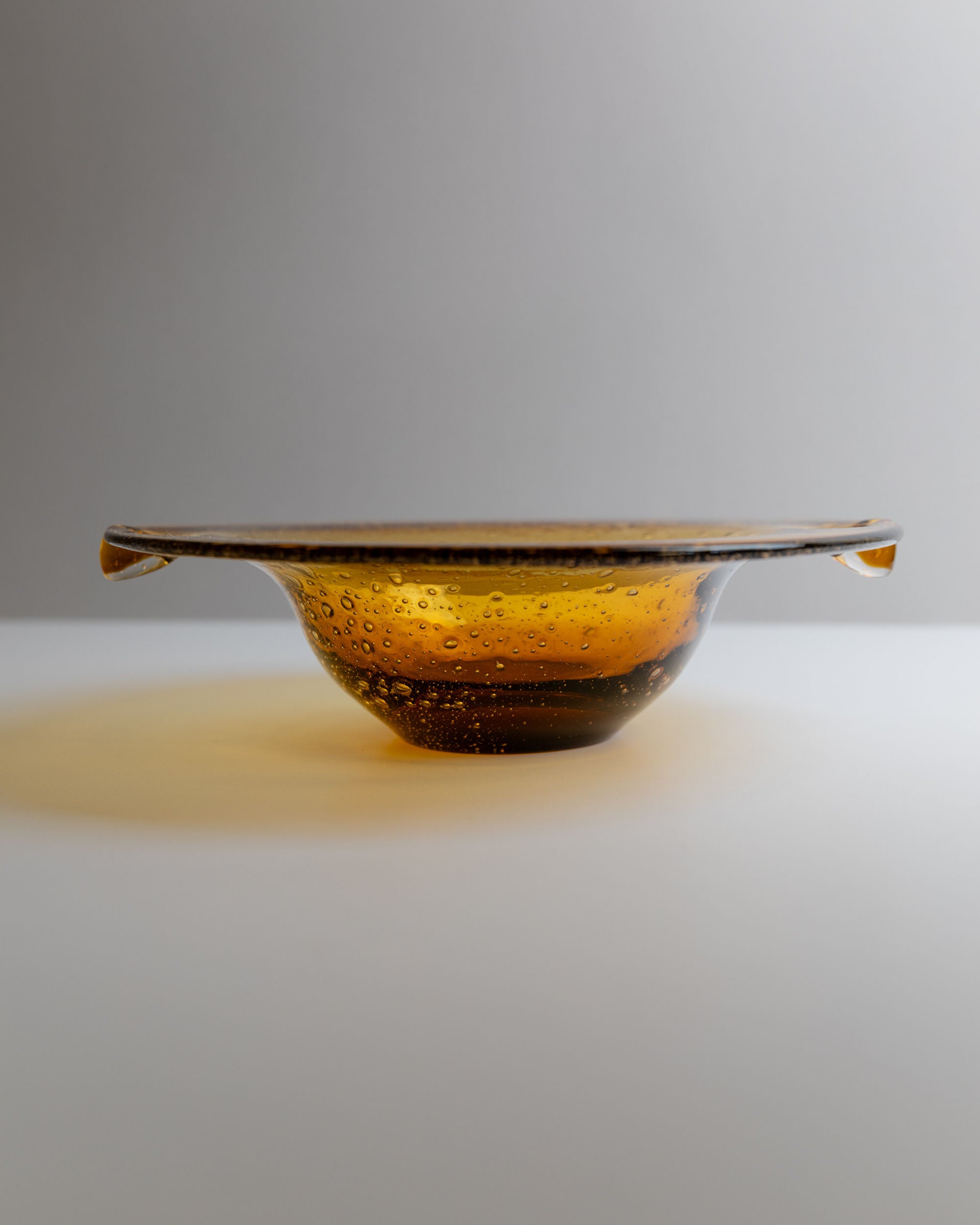 The Bottega Jacobs Murano Glass Bowl from Italy, 1970s, features a mid-century design with a slightly flared rim. Its amber hue casts a soft glow on the white surface, reminiscent of vintage Murano glass, adding decorative flair to the neutral backdrop with its charming brown bubbles.