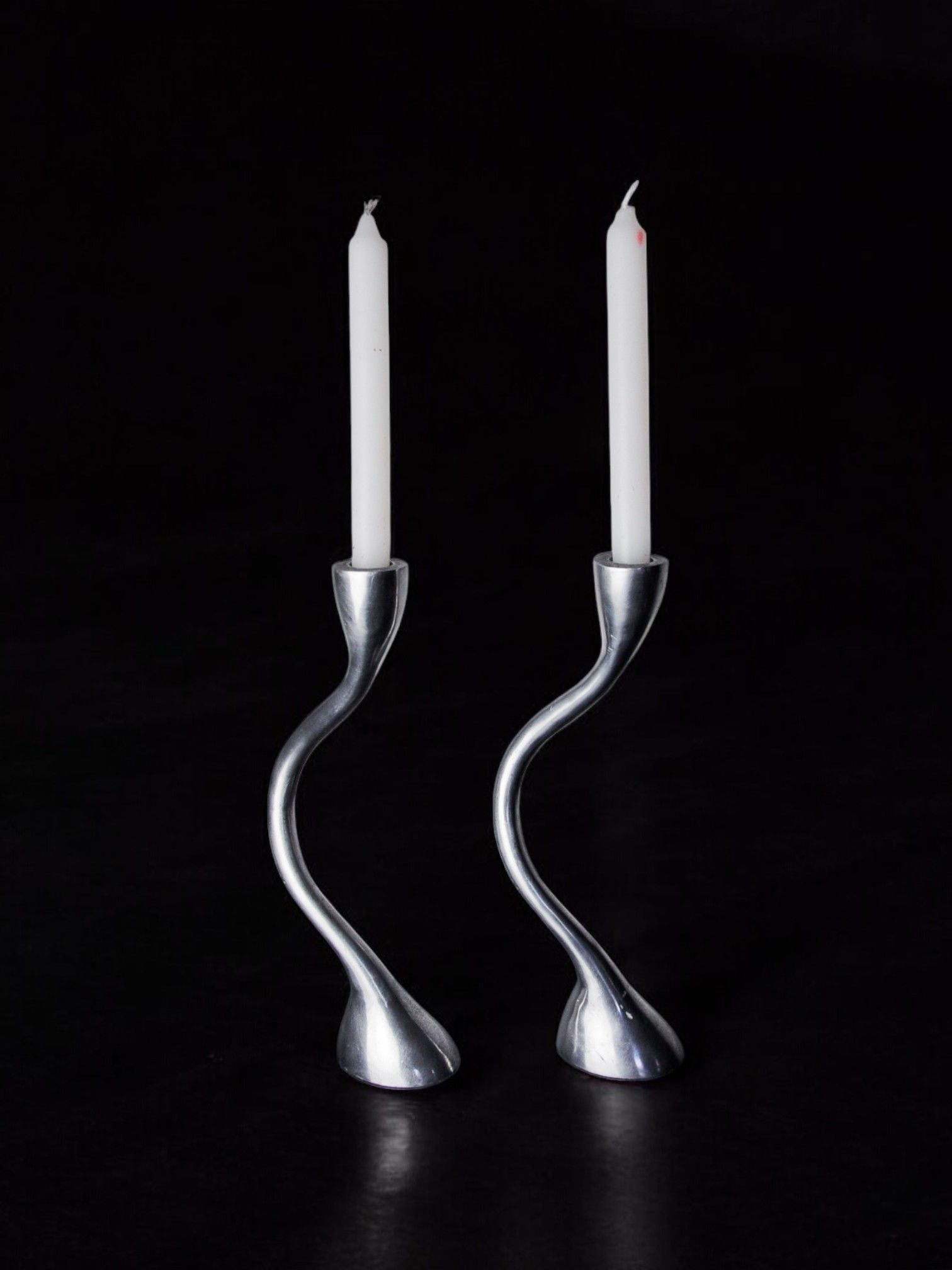 Two white candles in sleek, rare design objects from Malamar Studio's Pair of Zigzag Candle Holders 1970 set against a dark background.