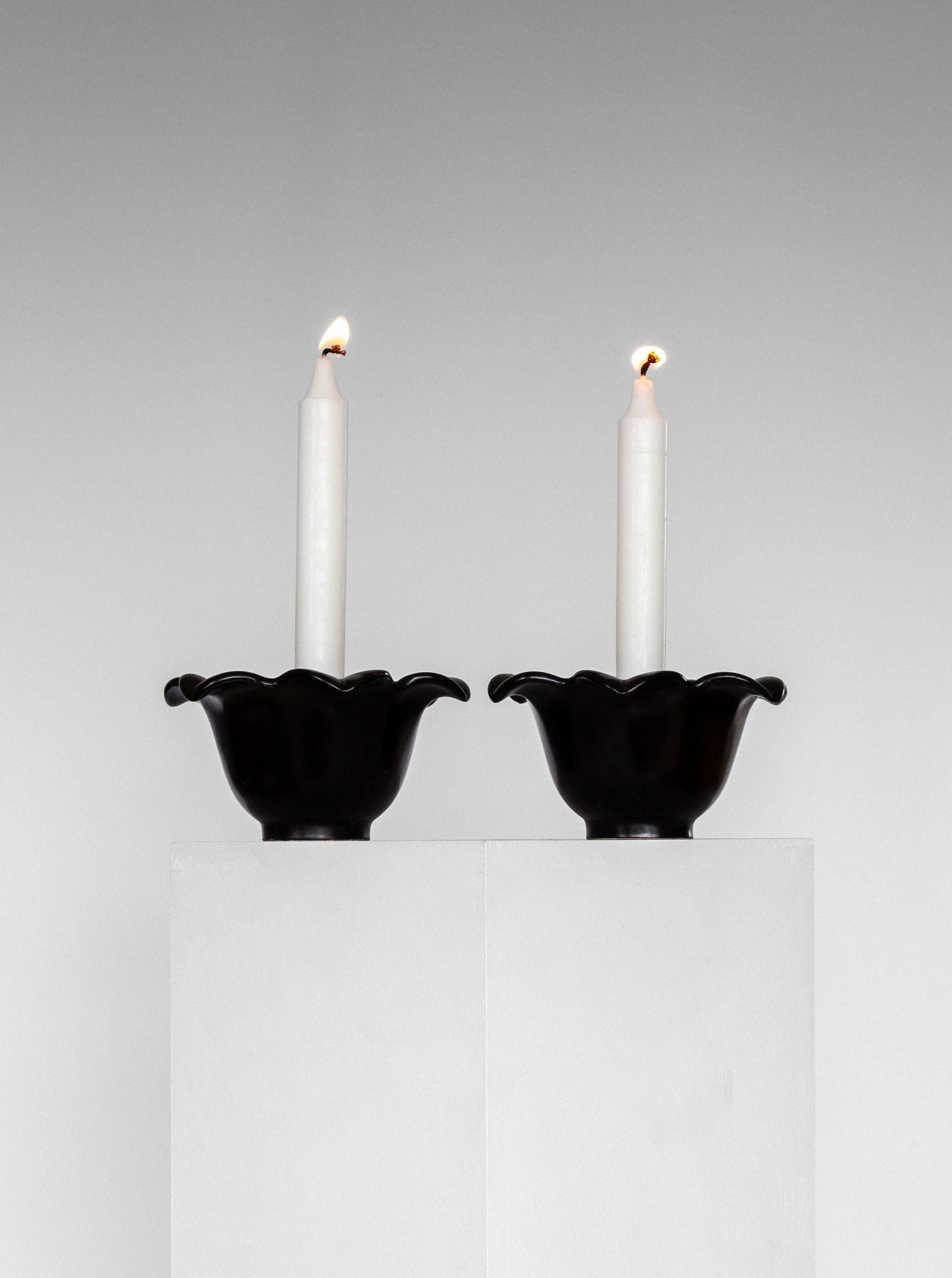 Two white candles are lit and placed in matching black, scallop-edged Spigel Mid-Century Ceramic Candlesticks from Sweden, 1950s. The candlesticks, reminiscent of Swedish Mid-Century Modern studio pottery by Lillemor Mannerheim, are set on adjacent white pedestals against a plain, light gray background, creating a minimalist and symmetrical composition.
